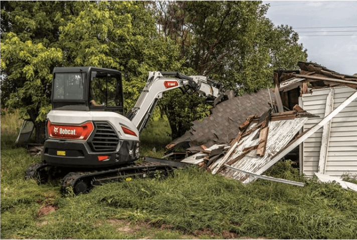Bobcat excavator demolishing a wooden house in a grassy area with trees in the background.