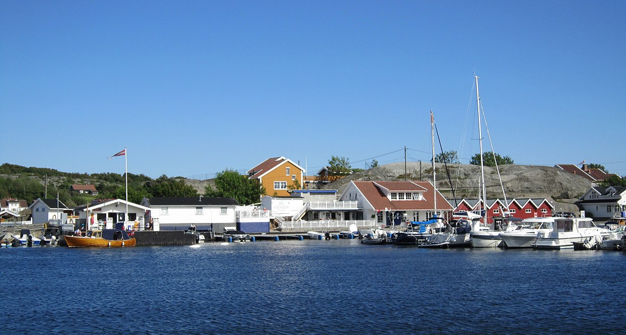Small marina with boats docked along the pier and colorful houses in the background under a clear blue sky.
