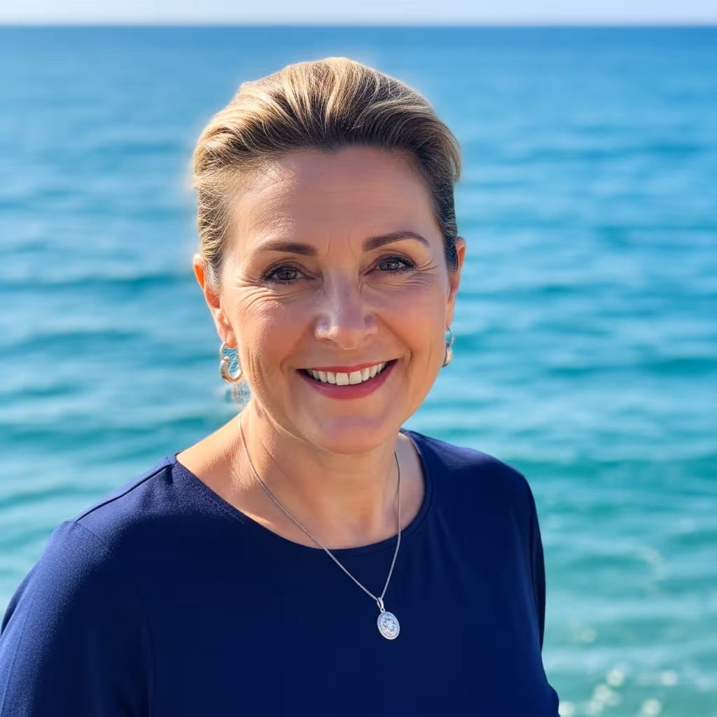 Smiling middle-aged woman with short blonde hair wearing a navy top and silver necklace by the ocean.