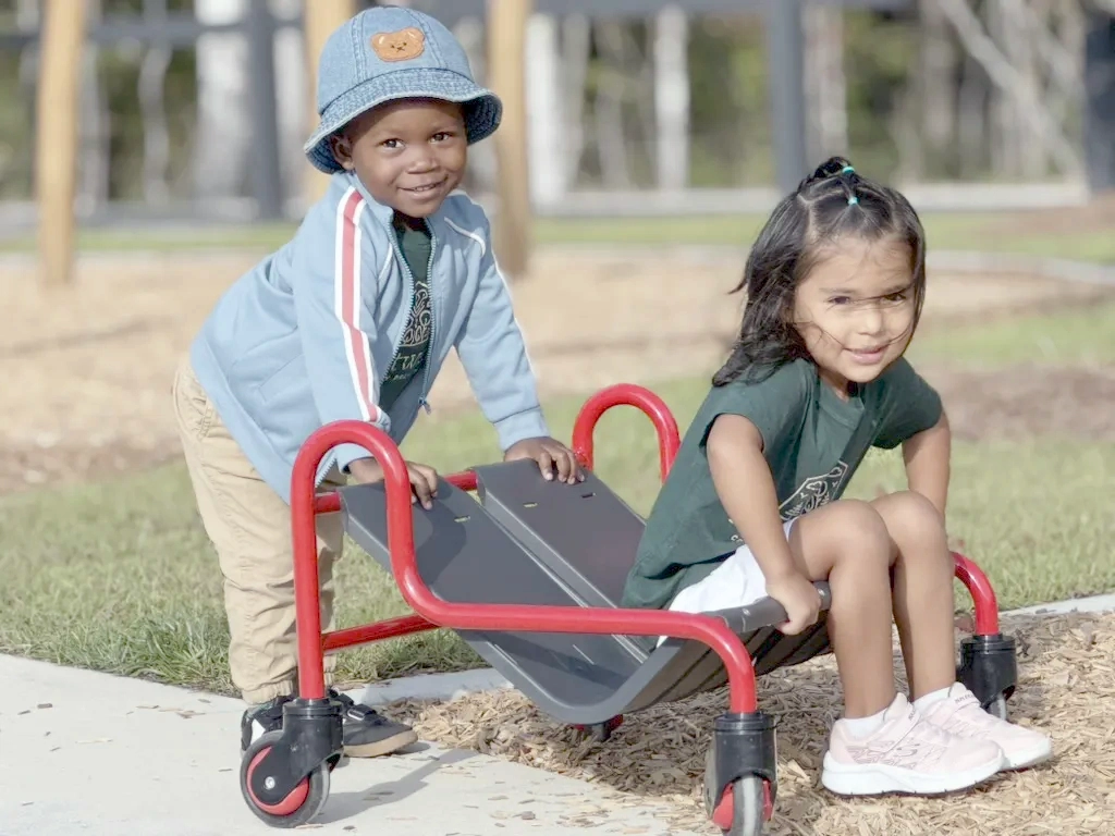 Two young children playing outdoors with a red and black wheeled scooter board on a sunny day.