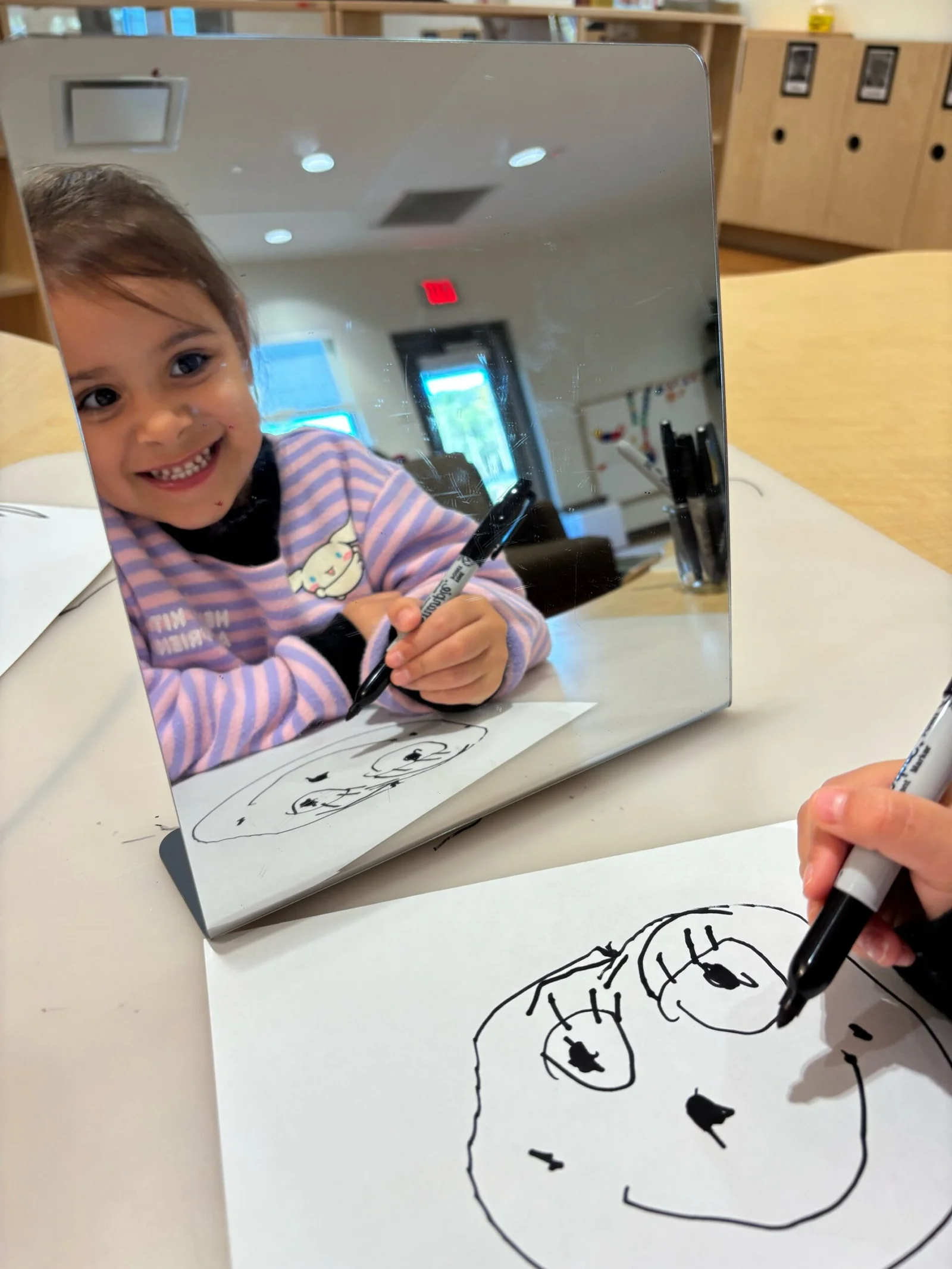 A young girl smiling while drawing a face with big eyes on paper, reflected in a mirror on a table.