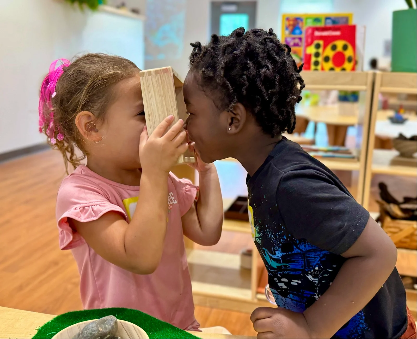 Two young children playing, one holding a wooden block to her eyes while the other looks closely at it.