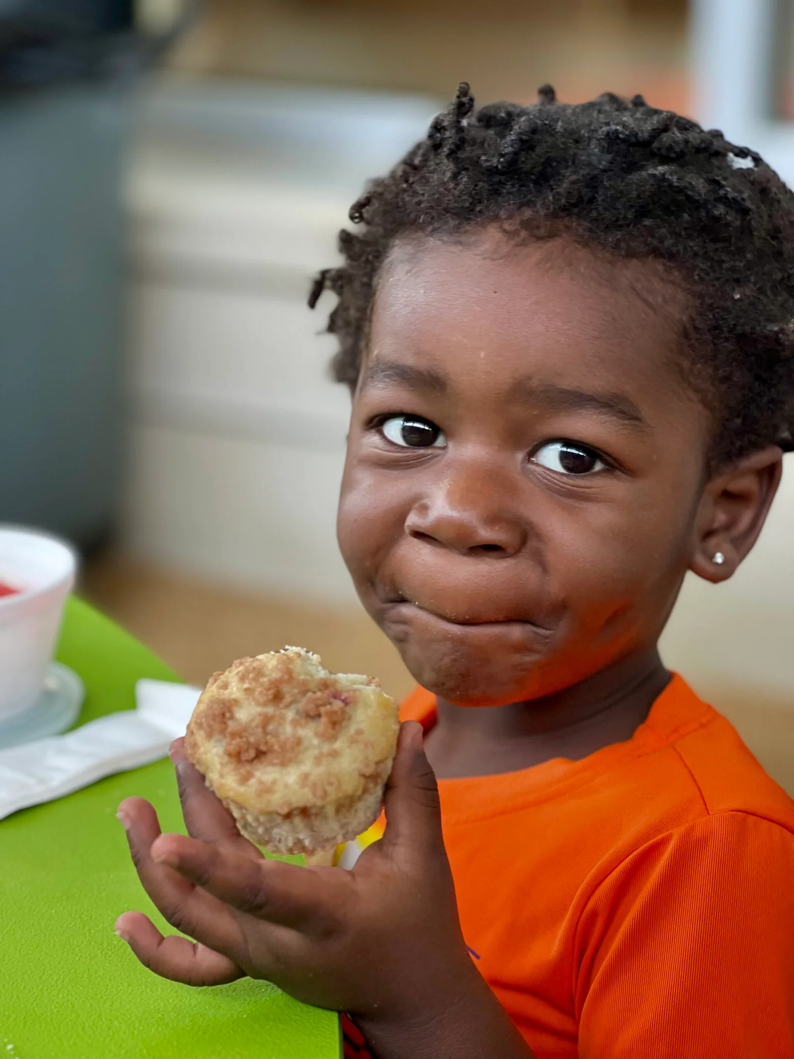 Young child in an orange shirt eating a crumbly muffin while sitting at a green table.