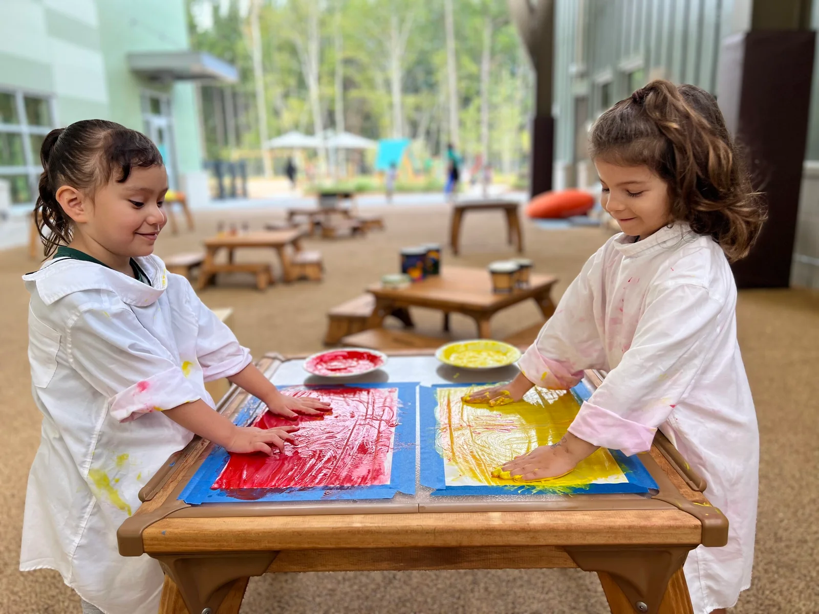 Two young girls wearing white smocks finger painting with red and yellow paint on a wooden table outdoors.