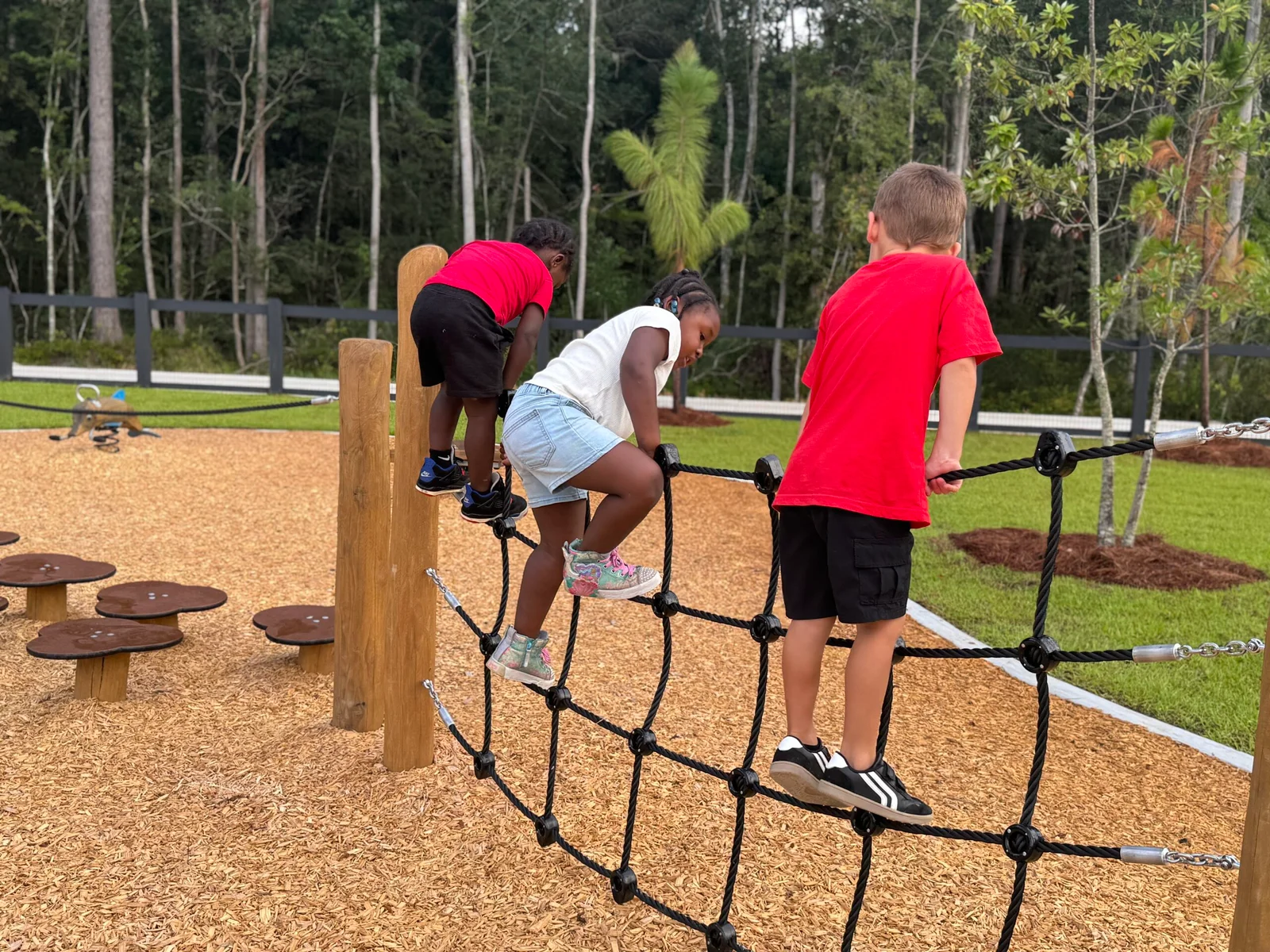 Three children climbing on a rope net structure at a playground with wood chip ground and trees in the background.