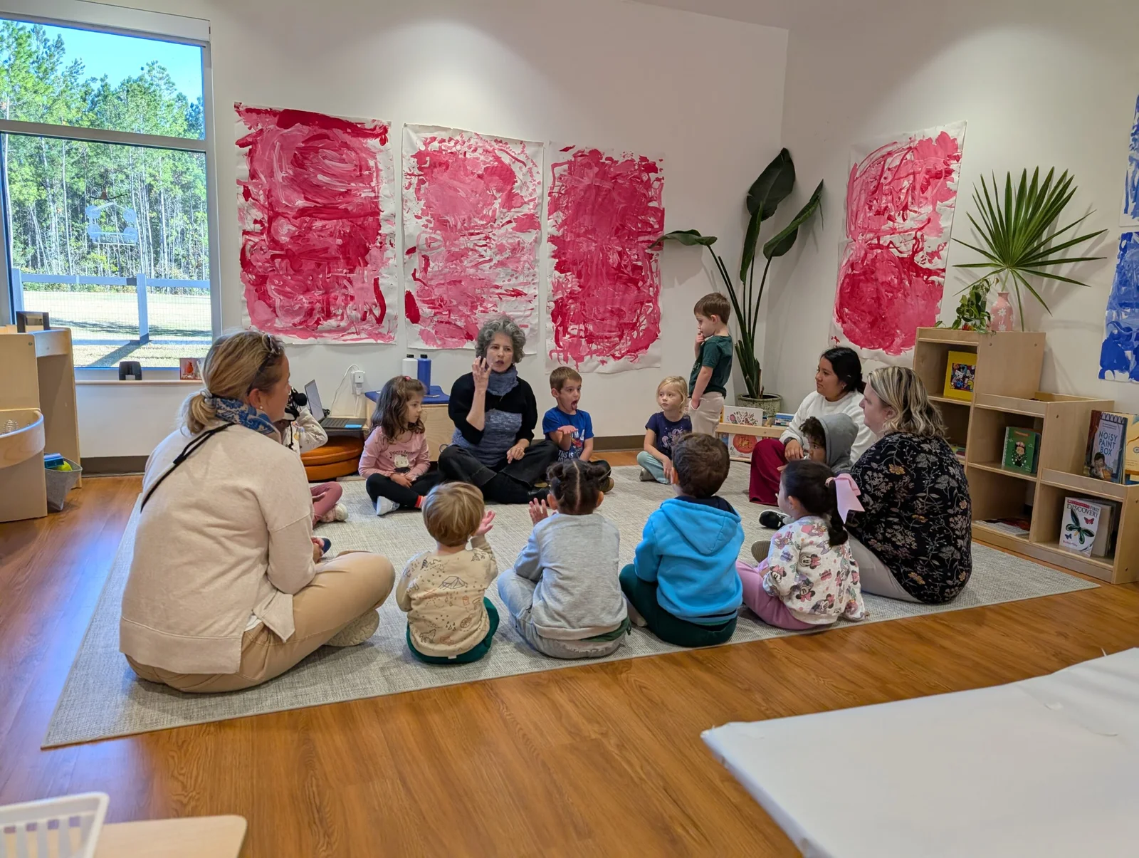 A group of children and two adults sit in a circle on a rug in a classroom with colorful red artwork on the wall.