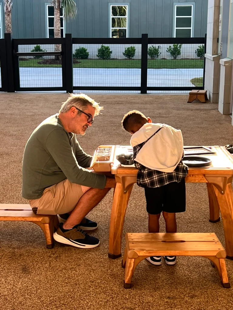Man wearing glasses sitting on a bench and interacting with a boy who is standing at a wooden table outdoors.