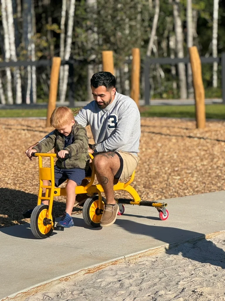Adult man and young boy riding a yellow tandem tricycle on a paved path at a playground.