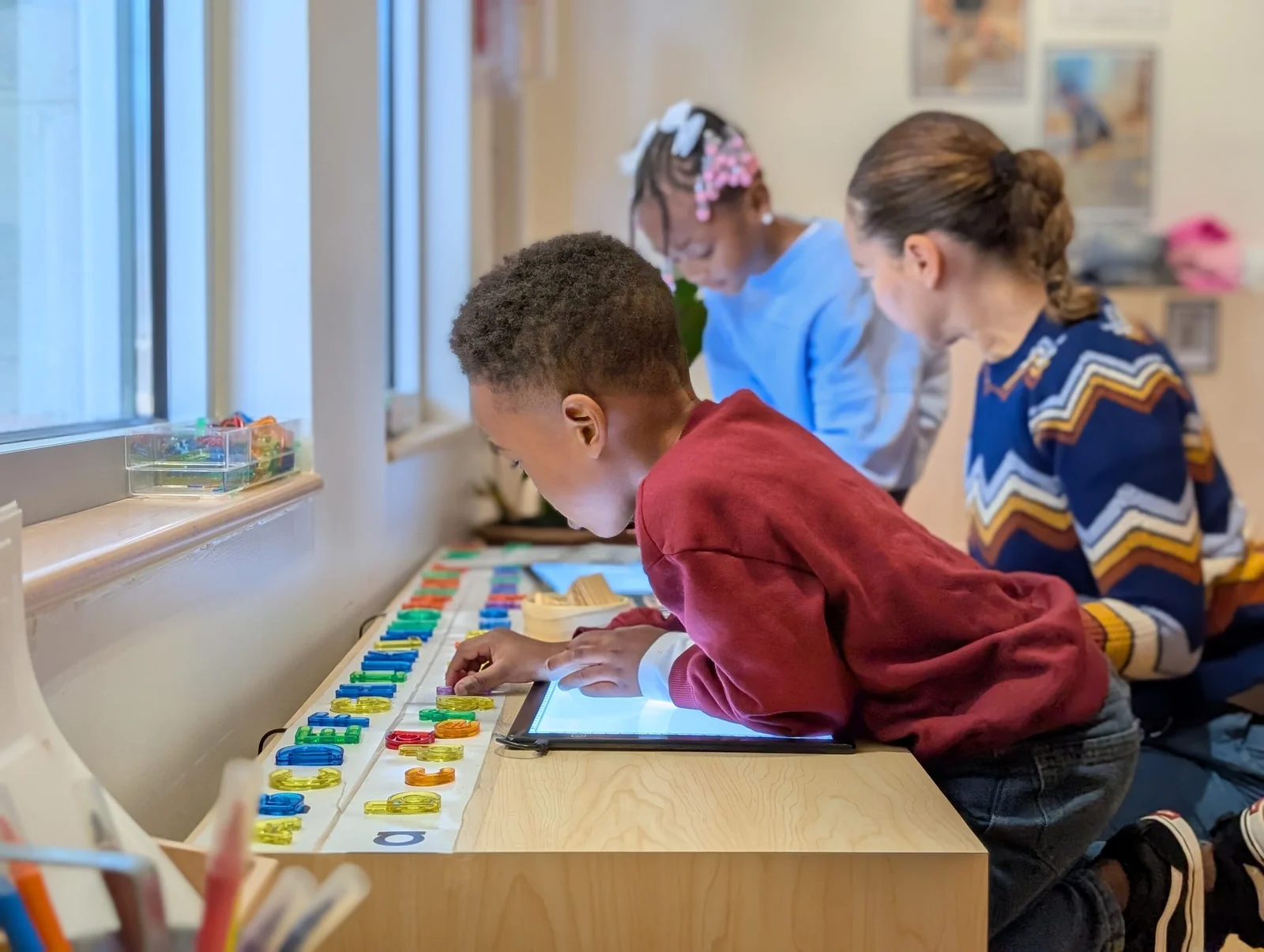 A child in a red sweater interacting with colorful translucent letters on a light table, with two other children and an adult nearby in a classroom.