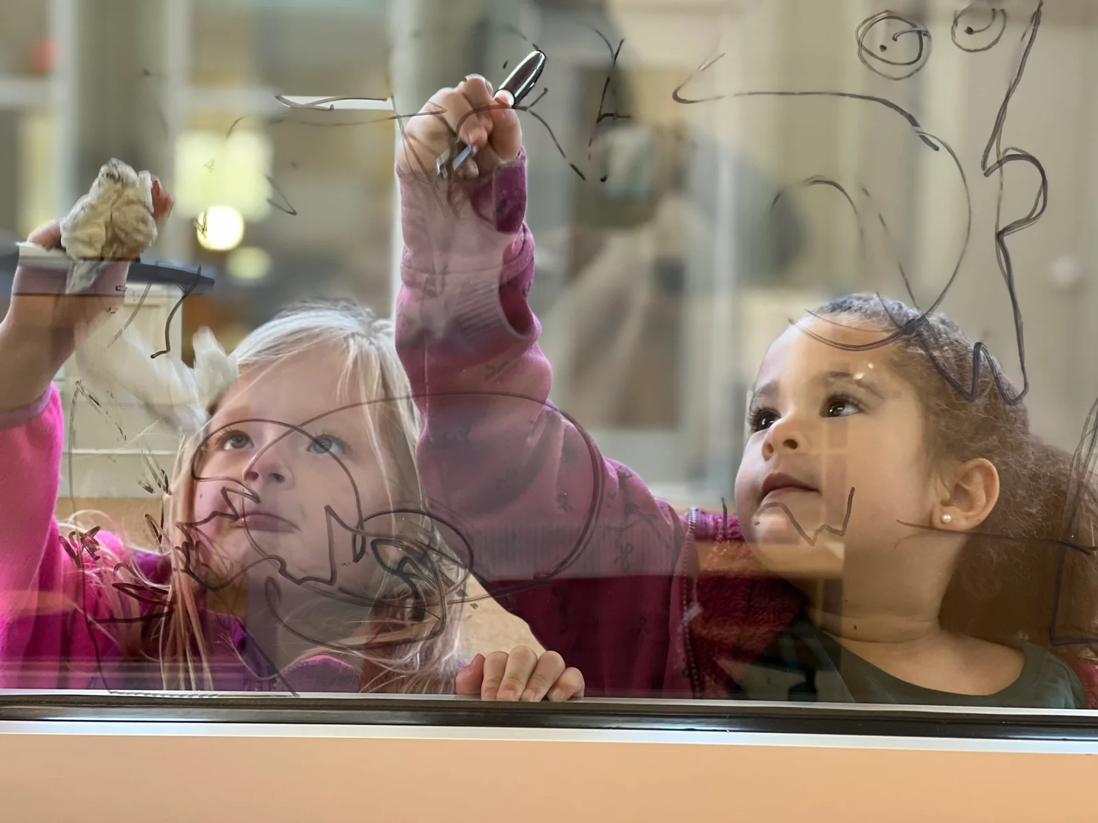 Two young children drawing on a clear glass window with markers, focused and engaged.