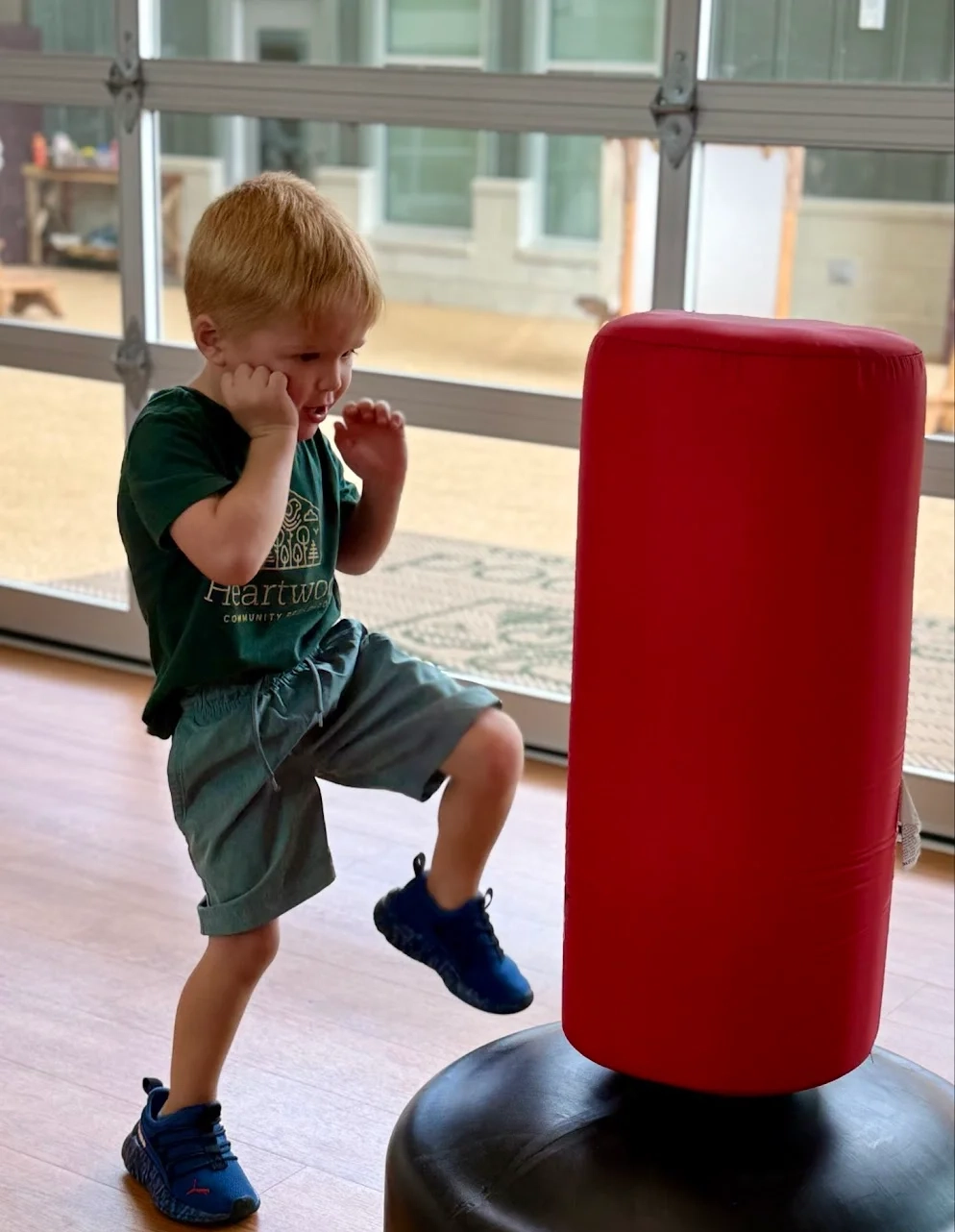 Young boy in blue shoes and green shirt practicing a knee strike on a red punching bag indoors.