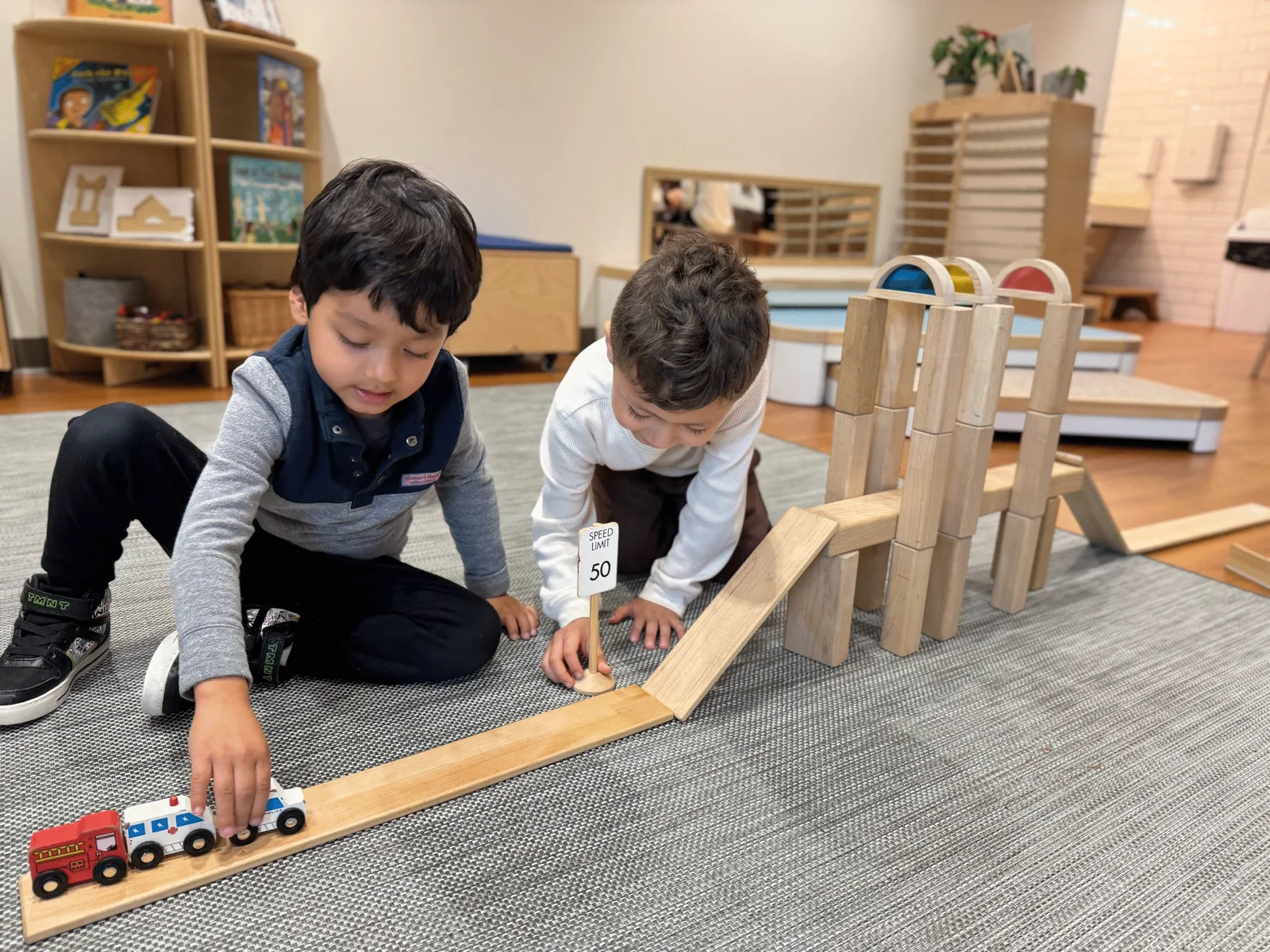 Two young boys playing on a gray carpet with a wooden ramp, toy vehicles, and a small speed limit sign inside a classroom.