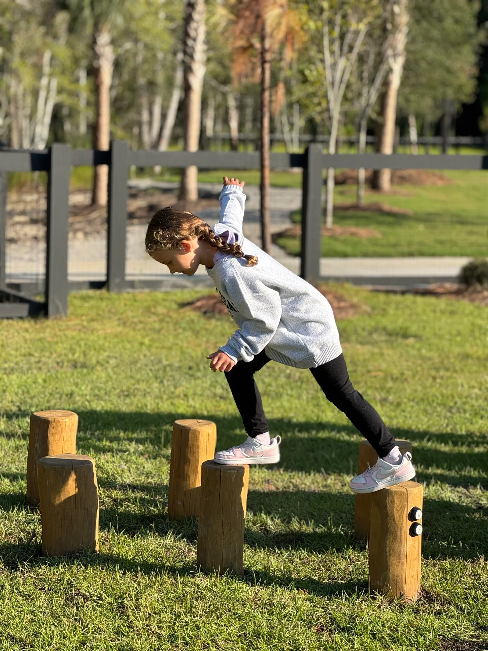 Child with a braided ponytail wearing a gray sweater and pink sneakers stepping across wooden posts in a grassy park.