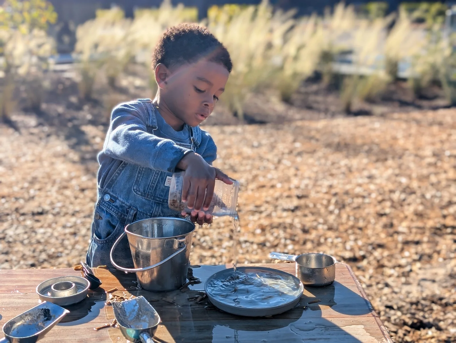 Young boy in denim overalls pouring water from a plastic cup into a metal bucket on a table outdoors.