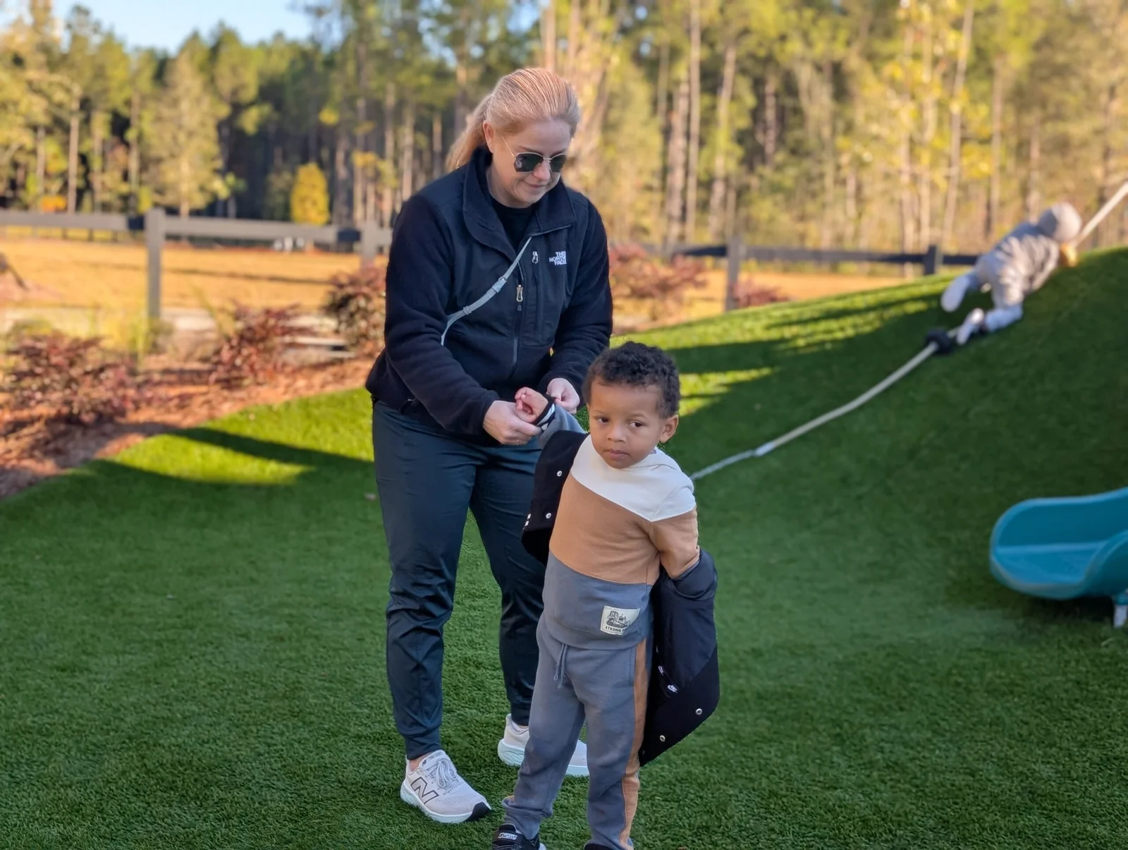 Adult helping a young child put on a jacket outdoors on green grass near a slide while another child plays on a grassy hill with a rope.
