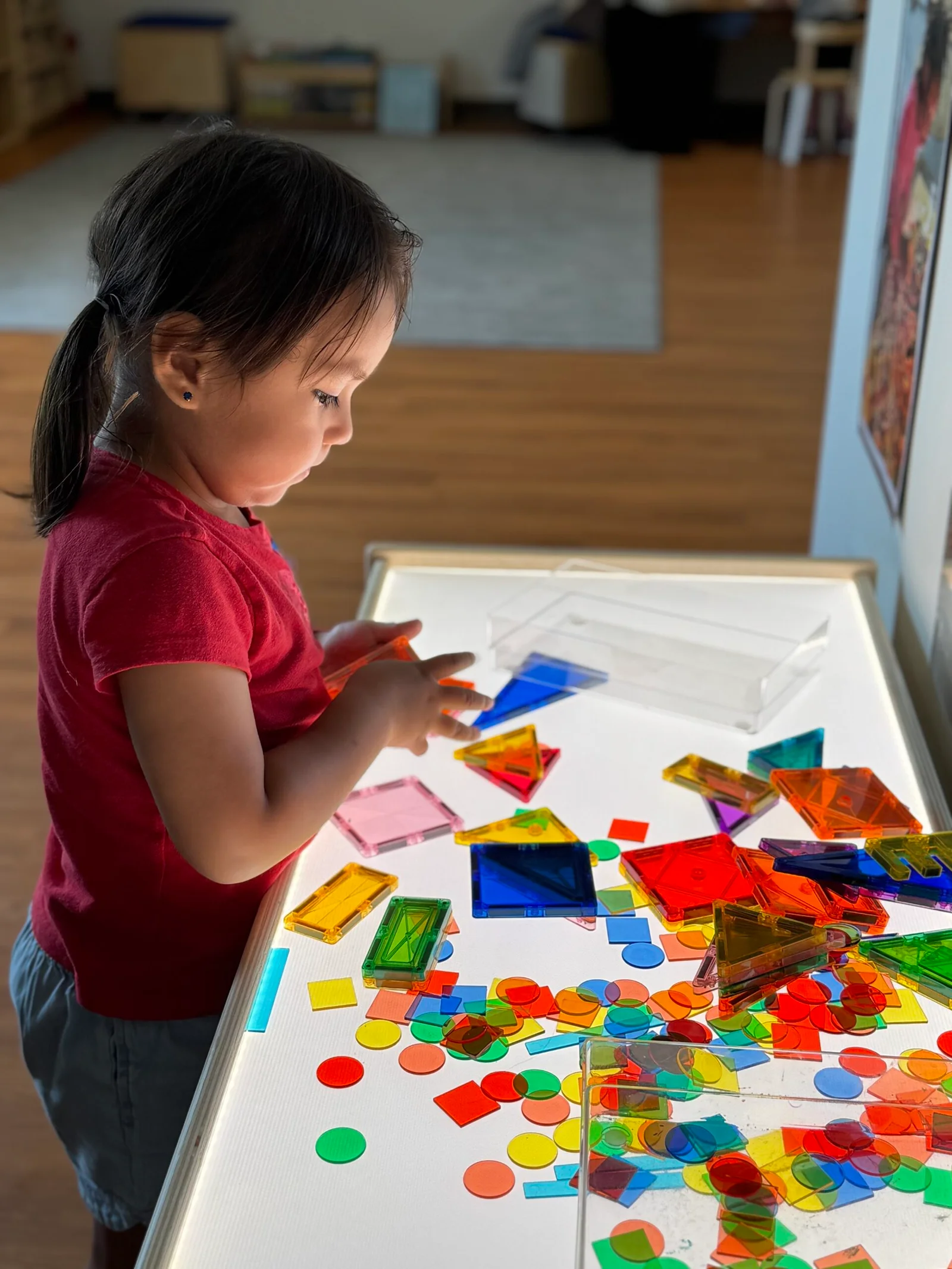 Young girl playing with colorful translucent shapes on a light table in a room with wooden floor.