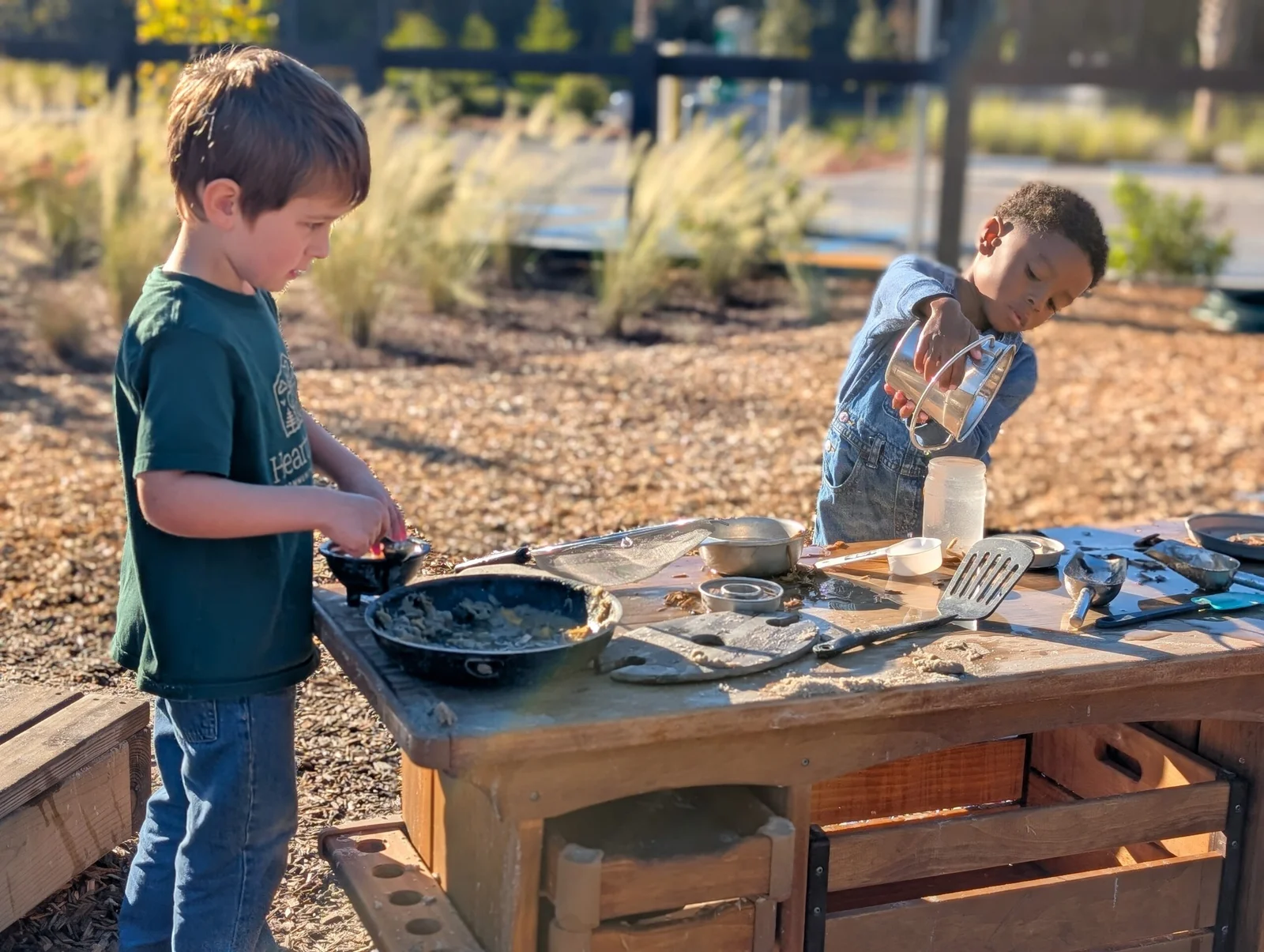 Two young boys playing with kitchen utensils and containers at an outdoor wooden play table.