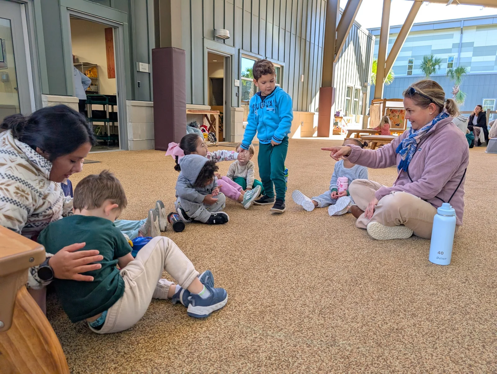 Children sitting in a circle on a carpeted floor with two adults, one pointing and engaging with them, in a bright indoor space.