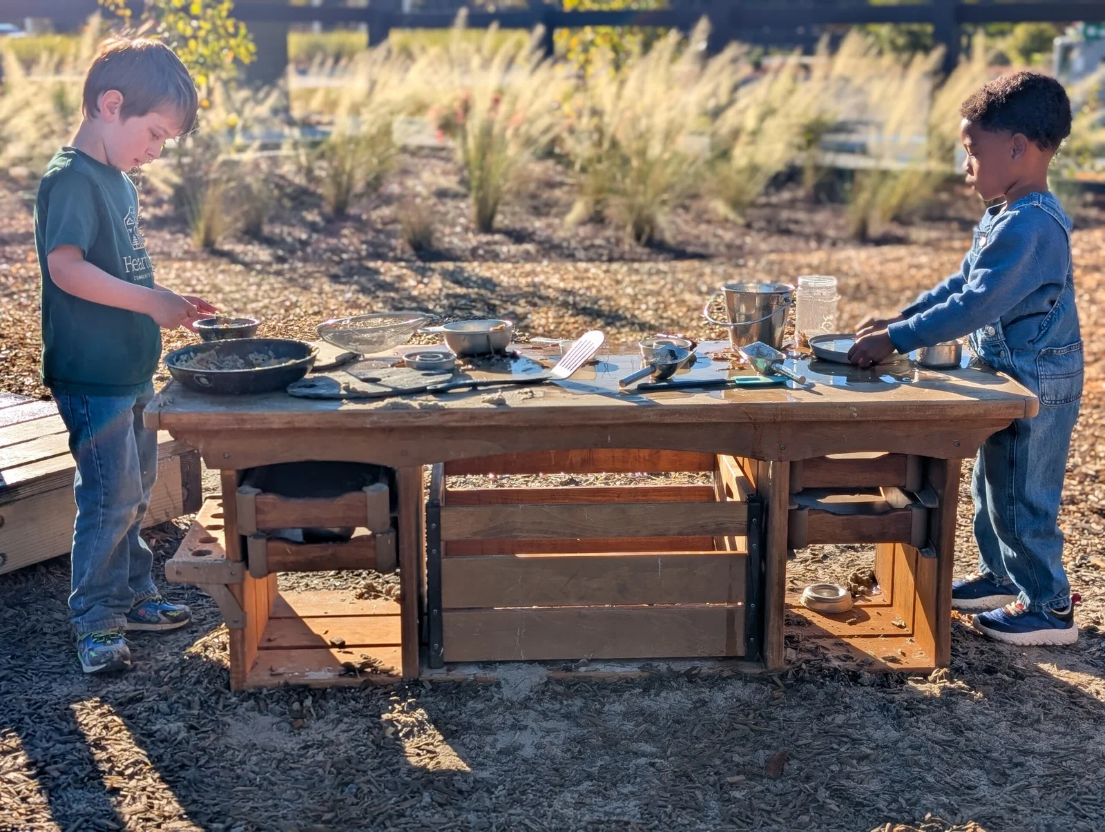 Two young boys standing at a wooden play table outdoors, engaged in sensory play with bowls, utensils, and containers on the table.