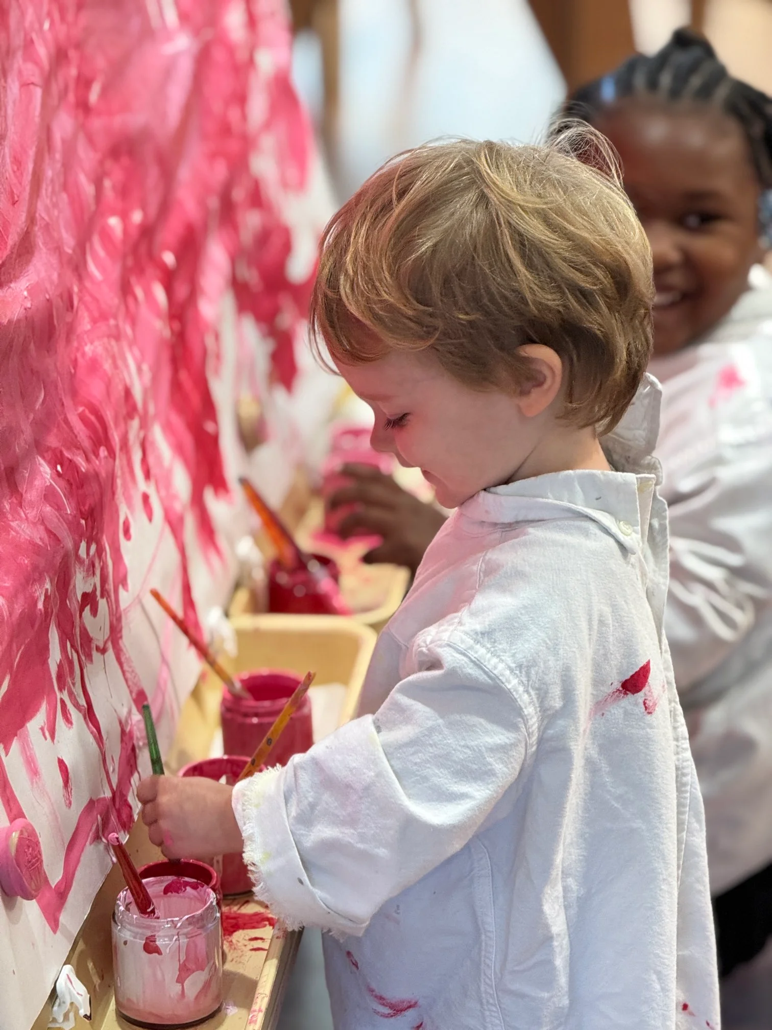 Young child in a white shirt painting with brushes dipped in pink and red paint on a wall, with another child smiling in the background.