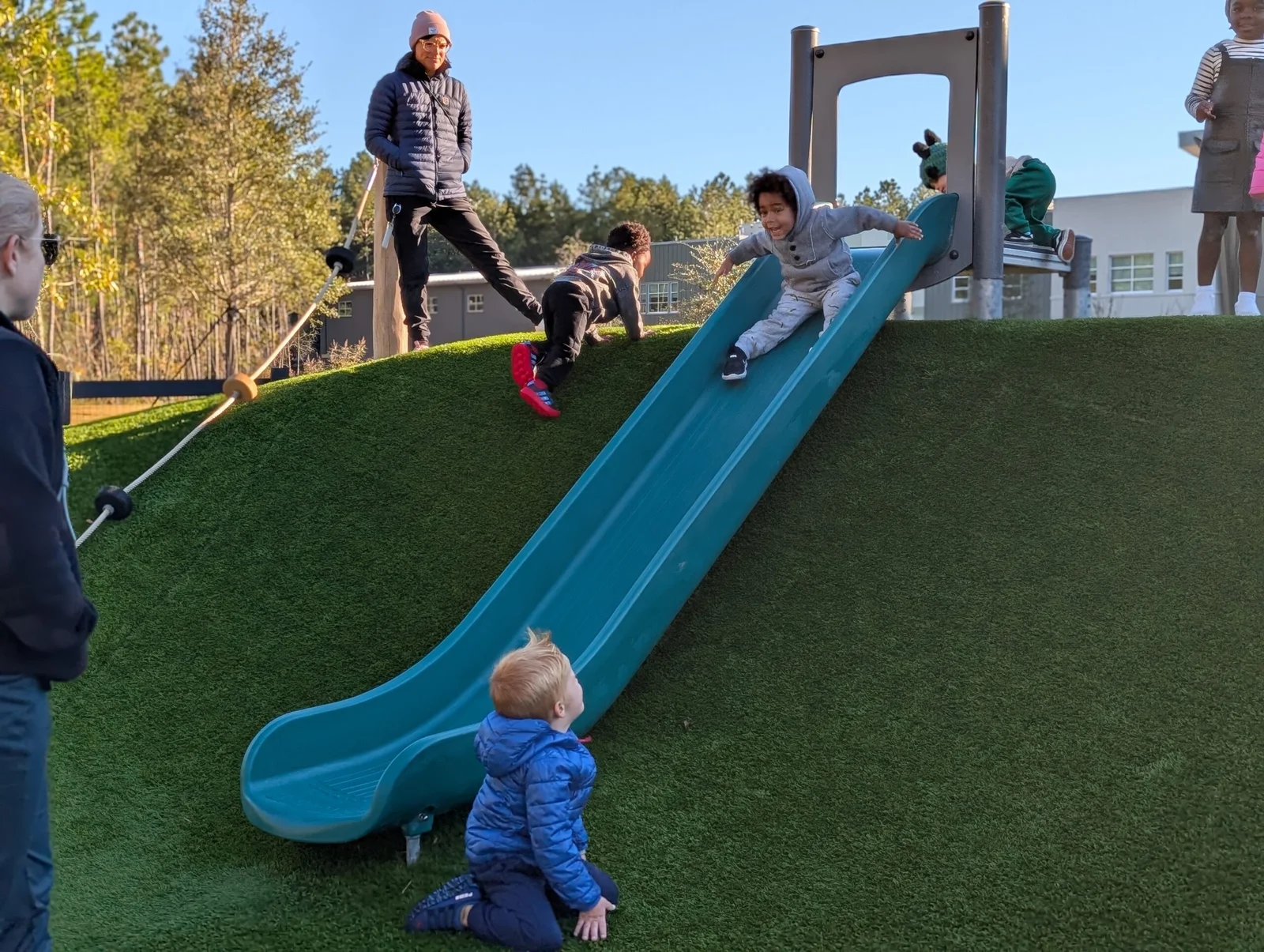 Children playing outdoors on a grassy hill with a blue slide under a clear sky, accompanied by adults.
