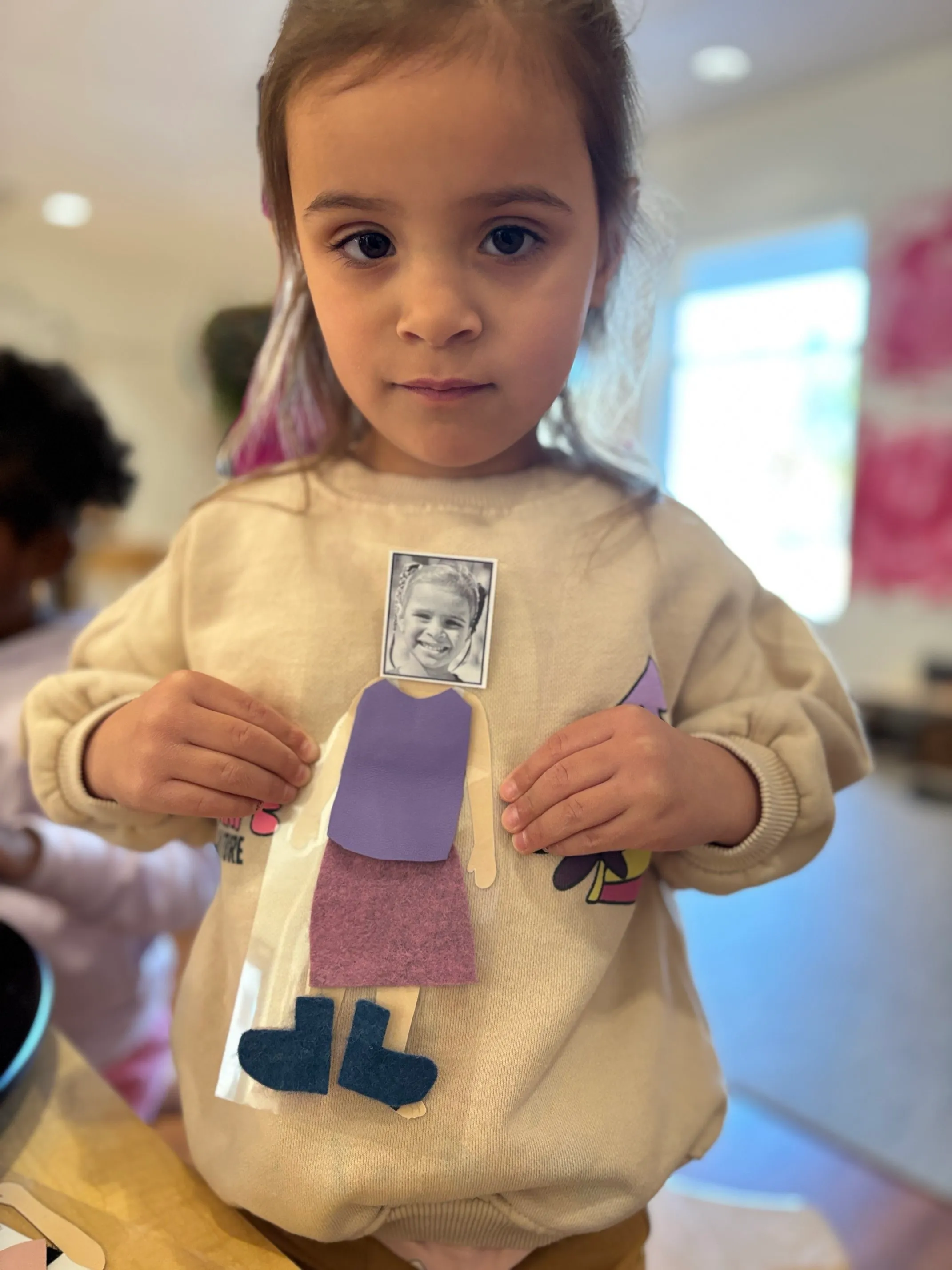 Young child holding a paper doll with a black-and-white photo of a smiling face, dressed in purple and blue paper clothes.