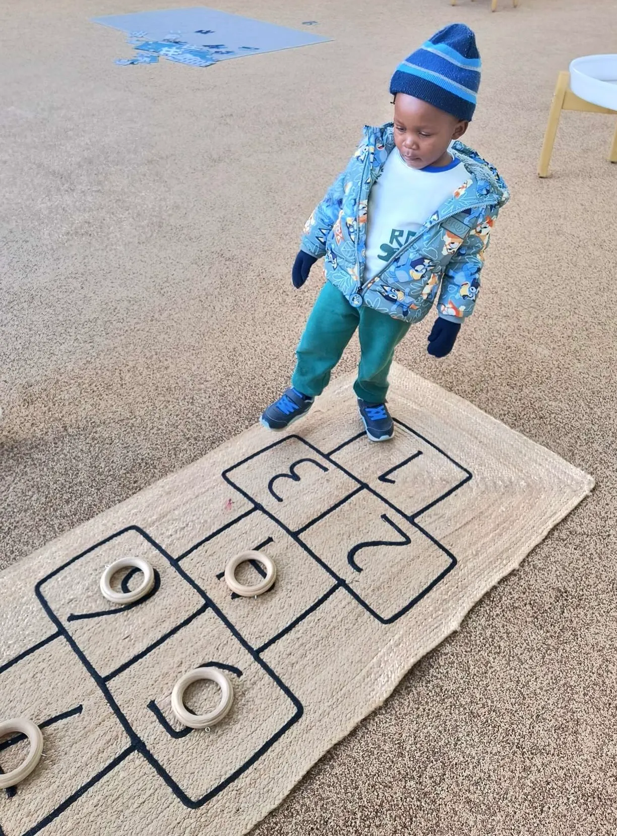 Child wearing a blue jacket, hat, and gloves standing on a numbered ring toss mat with wooden rings on some numbers.