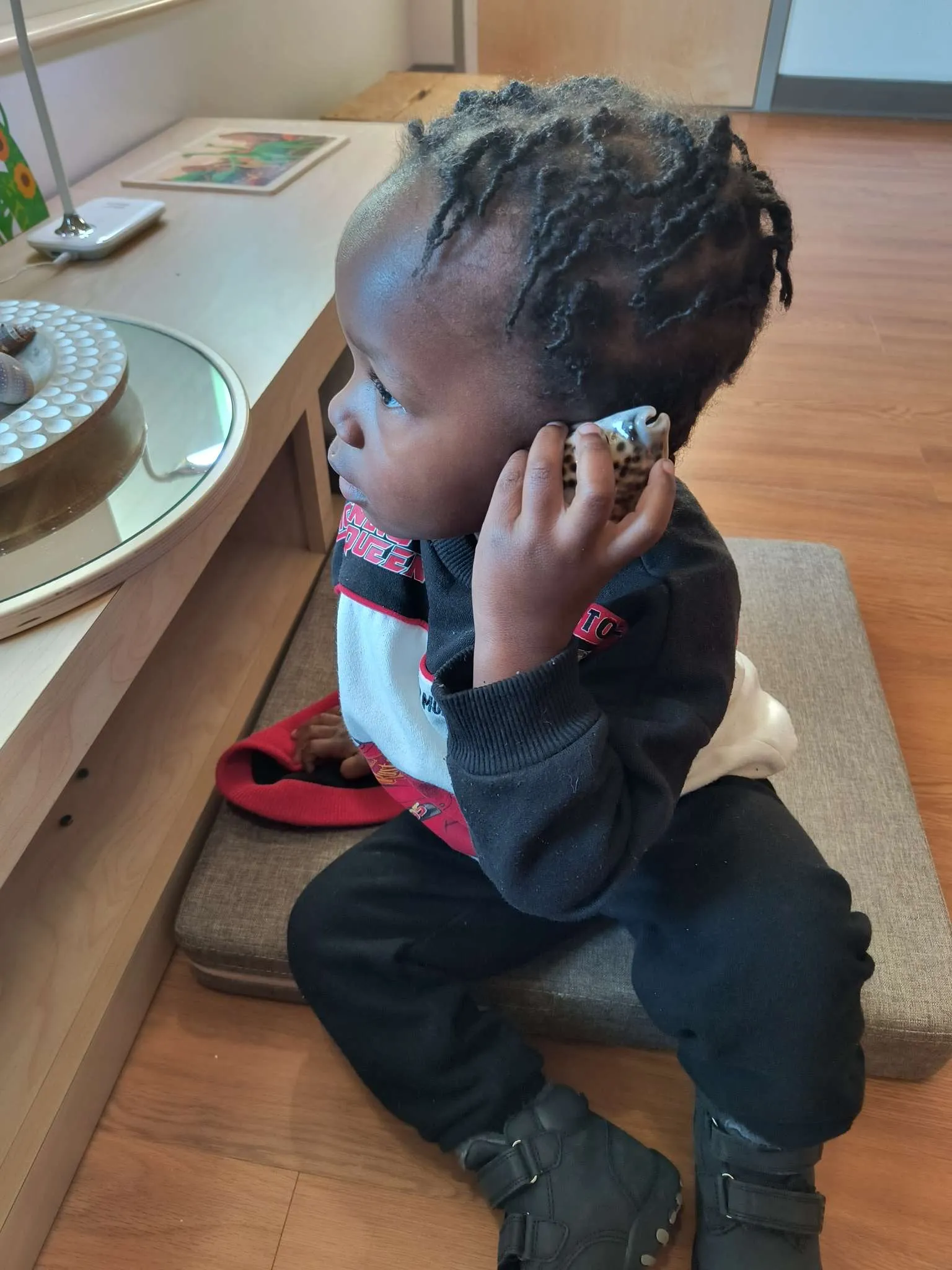 Young child with braided hair sitting indoors on a cushion holding a seashell to their ear.