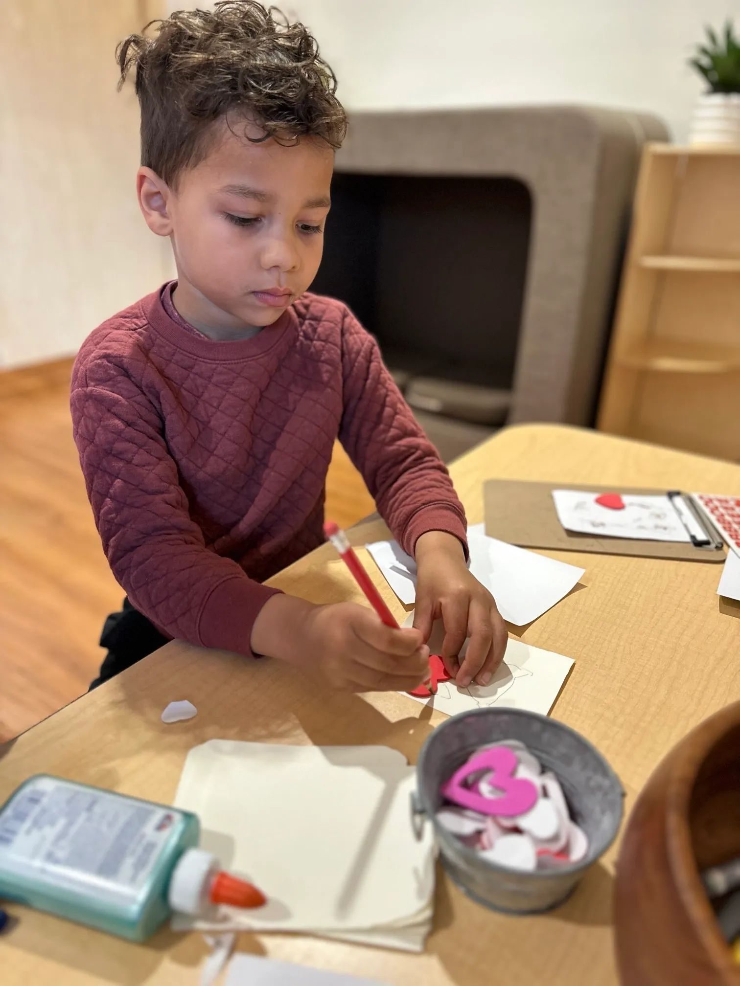 Young boy in a maroon sweater drawing on paper with heart-shaped cutouts on a table.