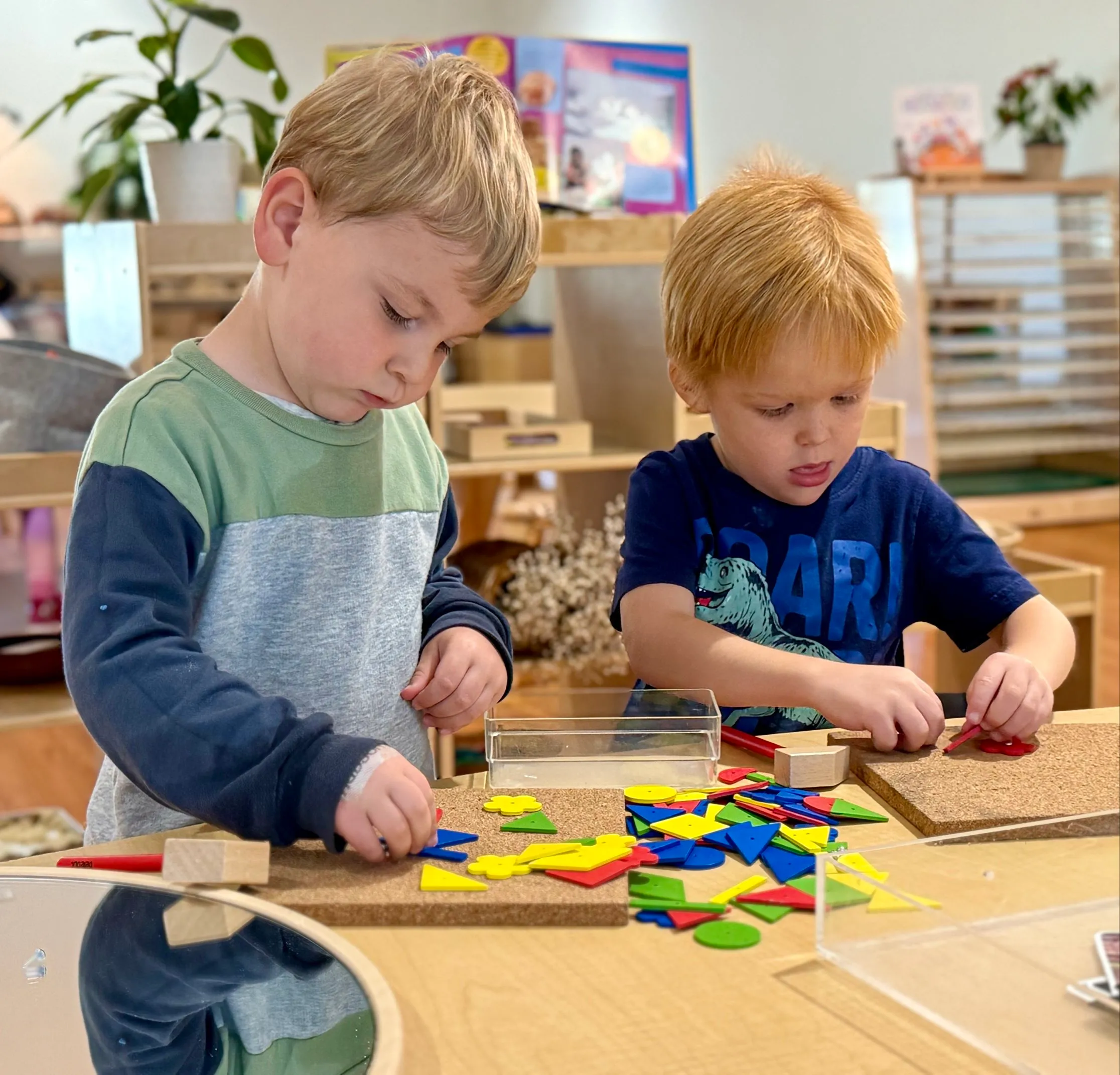 Two young boys focused on arranging colorful geometric shapes on cork boards at a wooden table in a classroom.