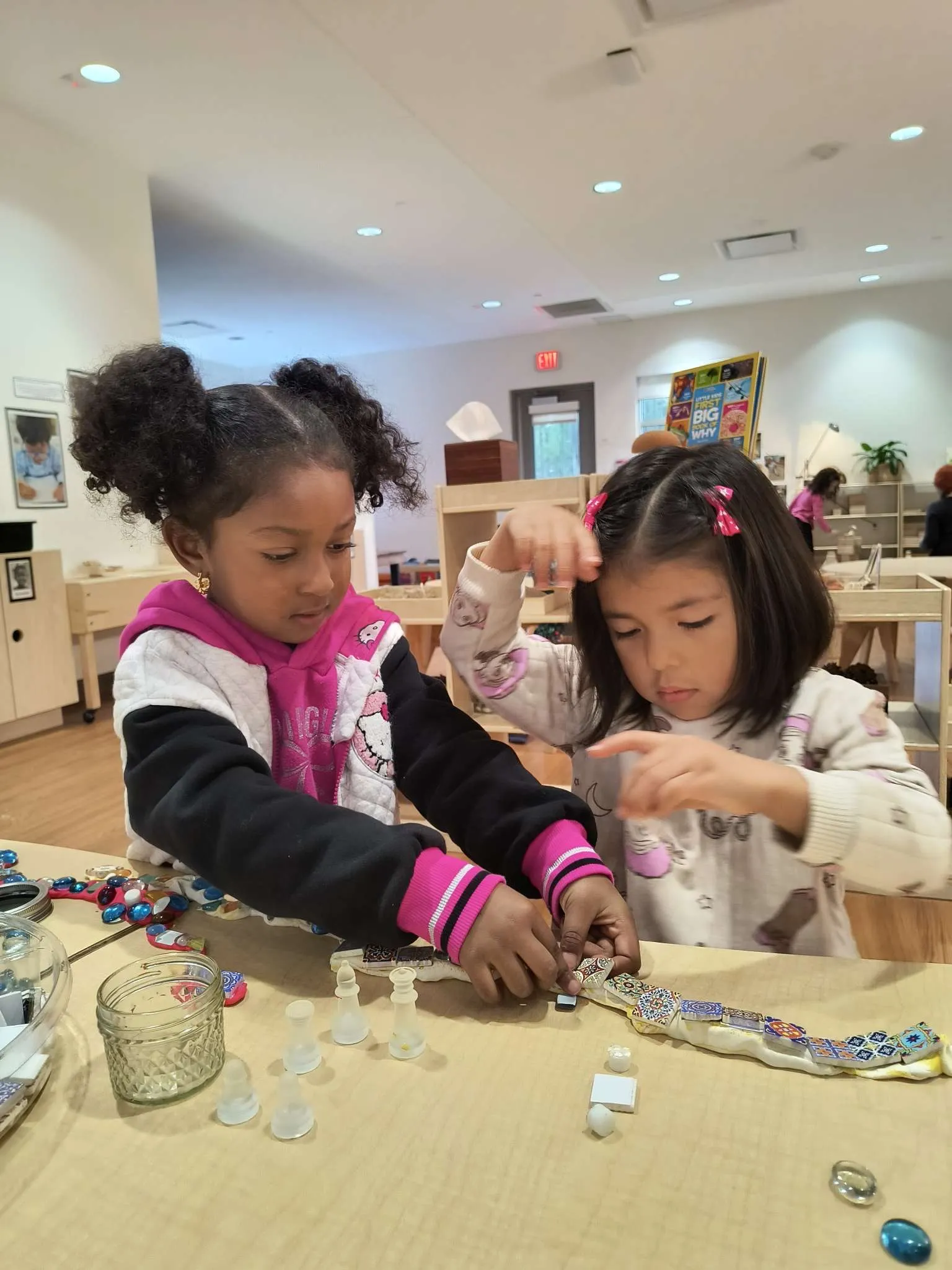 Two young girls focused on arranging colorful tiles and glass pieces on a table in a bright classroom.