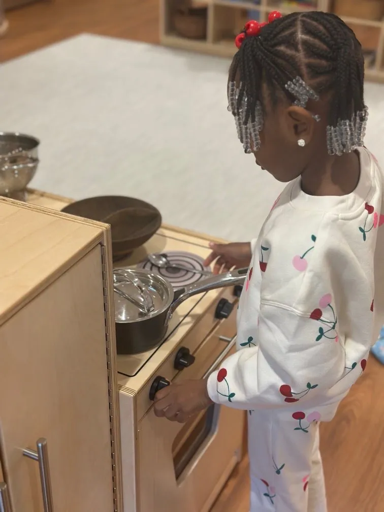 Young girl with braided hair adorned with beads, wearing a white outfit with cherry patterns, is turning the knob of a wooden play kitchen stove.