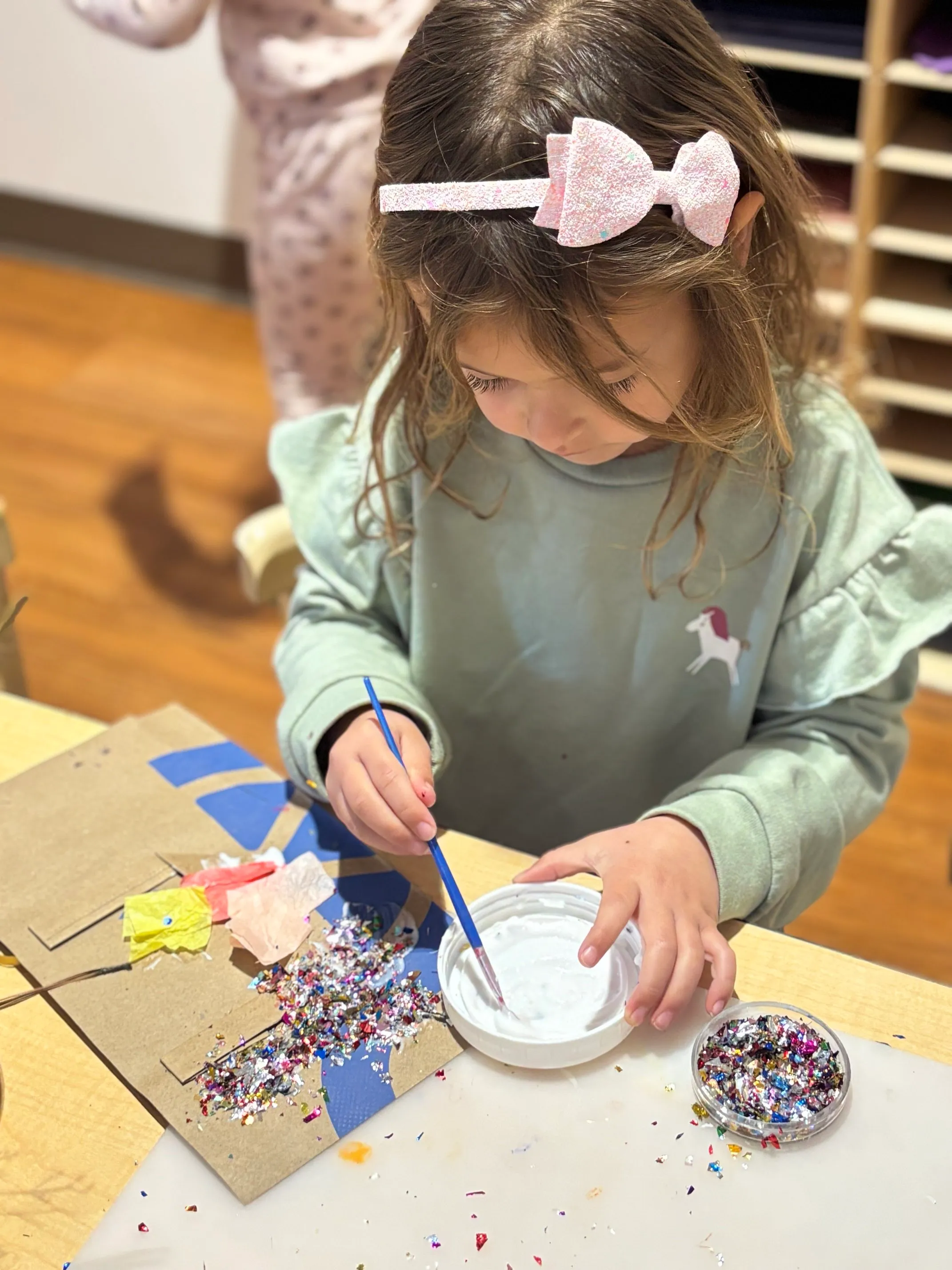 Young girl with a pink glittery bow headband painting with a brush while doing a craft with colorful glitter and tissue paper on a table.