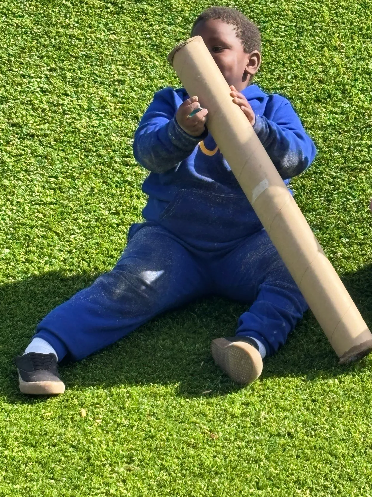 Young child sitting on green grass playing with a large cardboard tube.