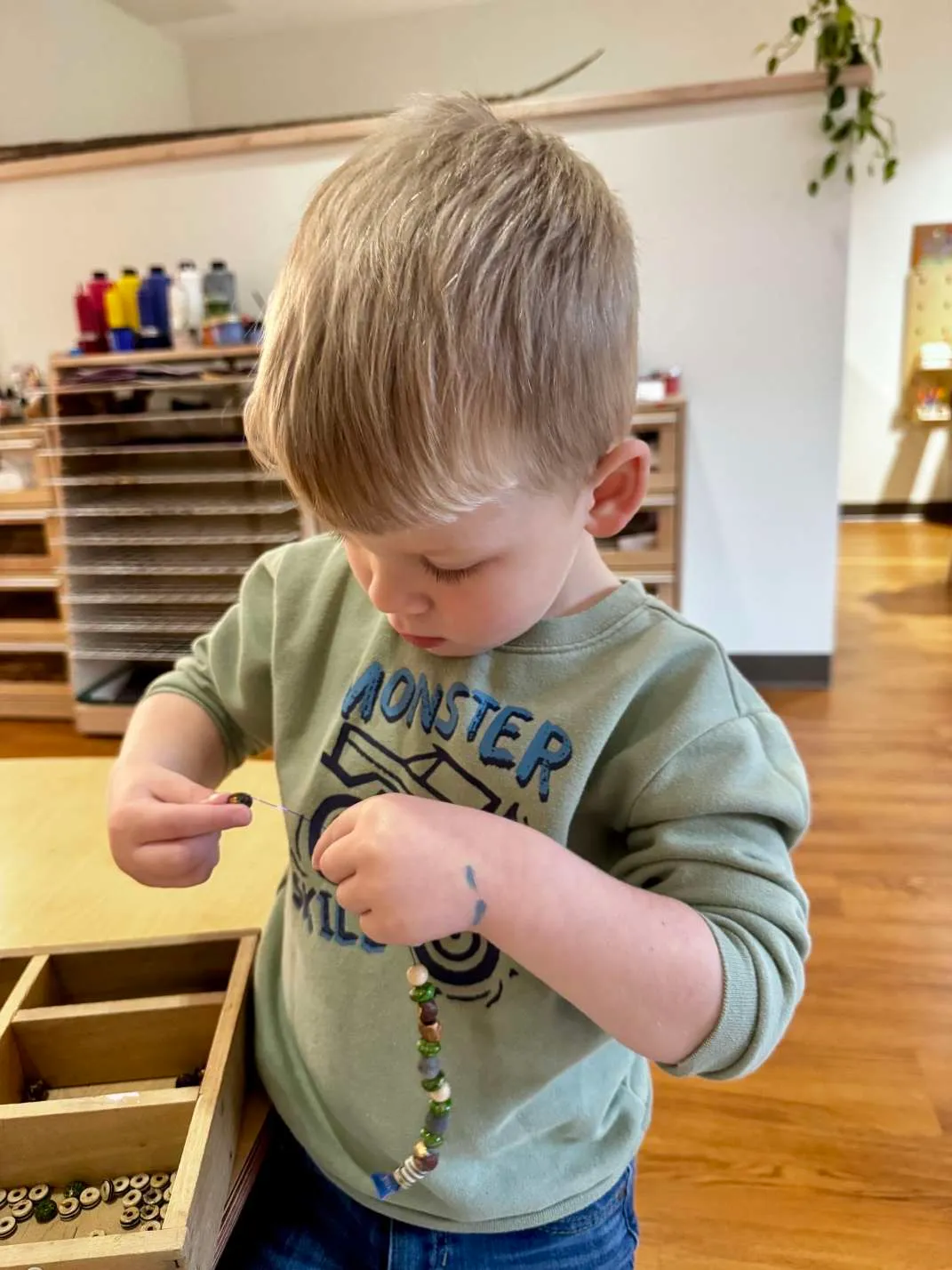 Young boy threading beads onto a string while standing in a classroom with wooden flooring.