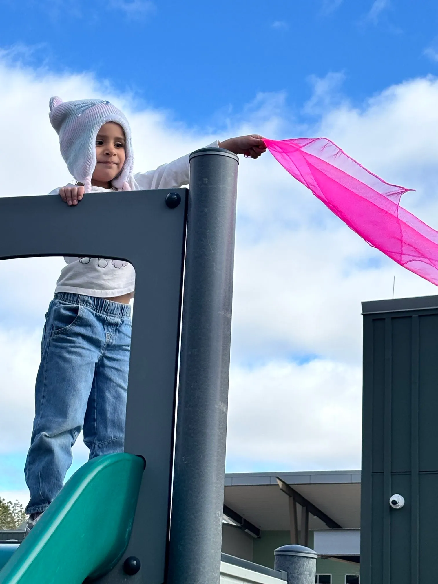 Child wearing a knit hat stands on playground equipment holding a flowing pink scarf against a blue sky with clouds.