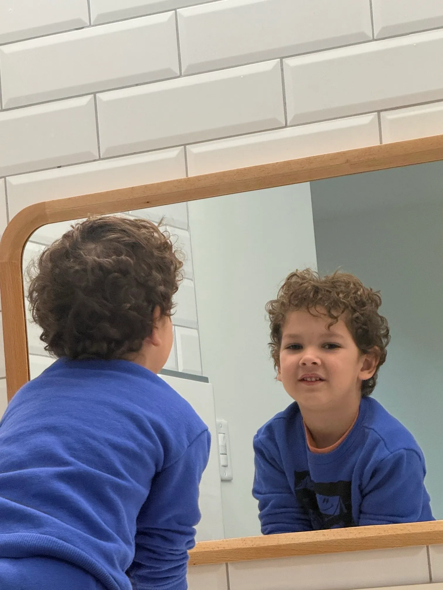 Young boy with curly hair wearing a blue sweater looking at his reflection in a rectangular wooden-framed mirror.
