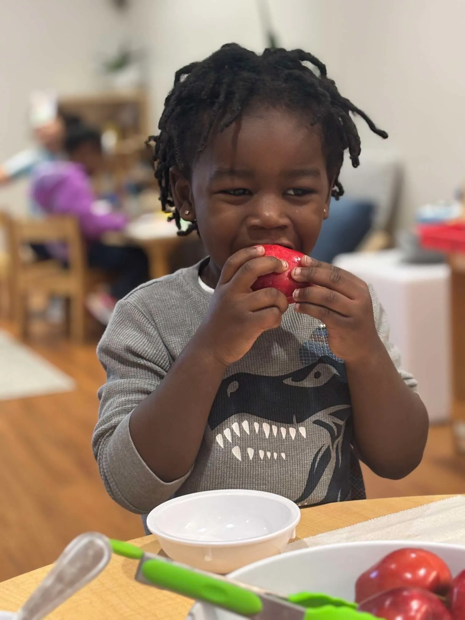 Child with braided hair wearing a dinosaur shirt bites into a red apple while sitting at a table with a white bowl and cutlery.