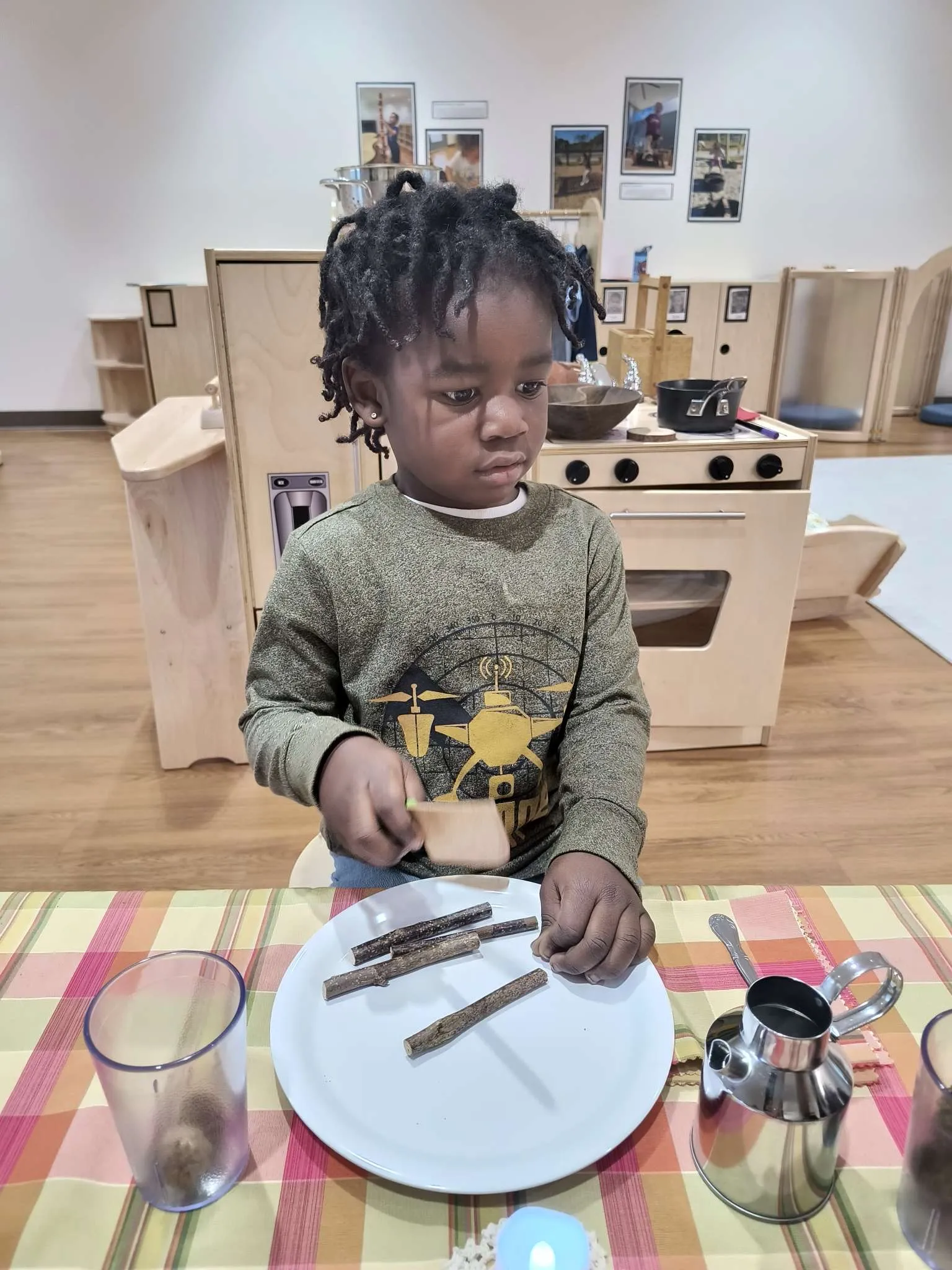 Young child seated at a table, playing with wooden sticks and holding a small wooden spatula in a classroom setting.