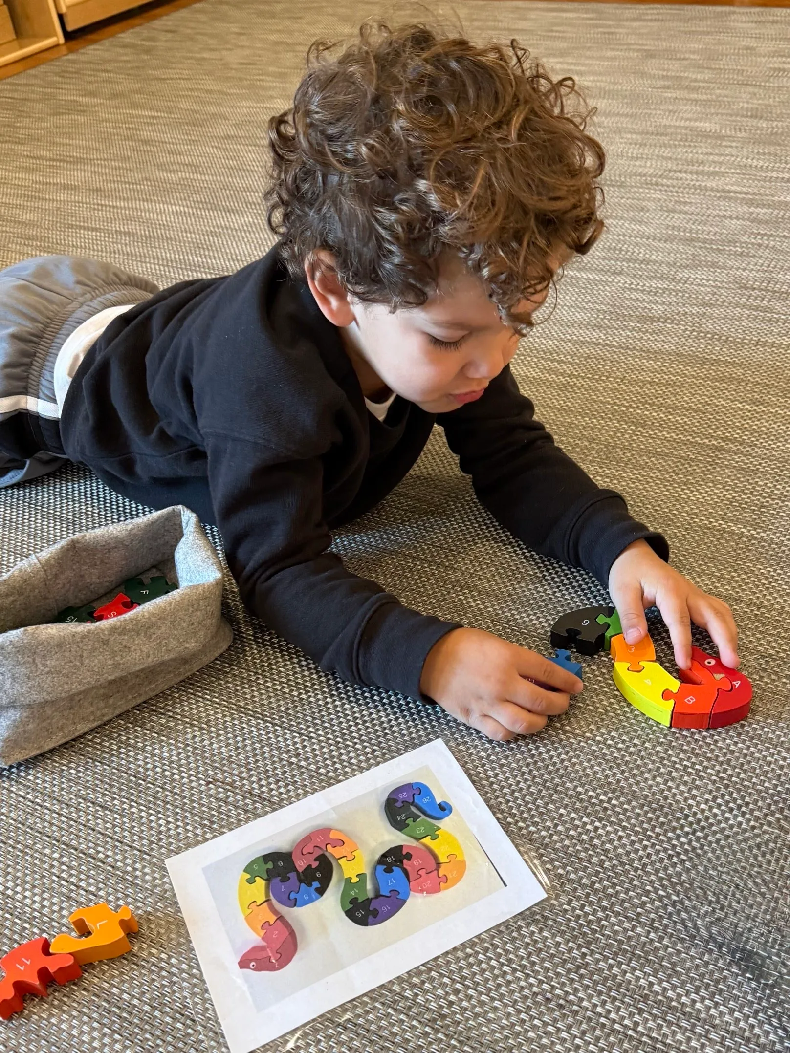 A young boy with curly hair lying on a textured carpet, assembling a colorful numbered puzzle with a picture guide beside him.