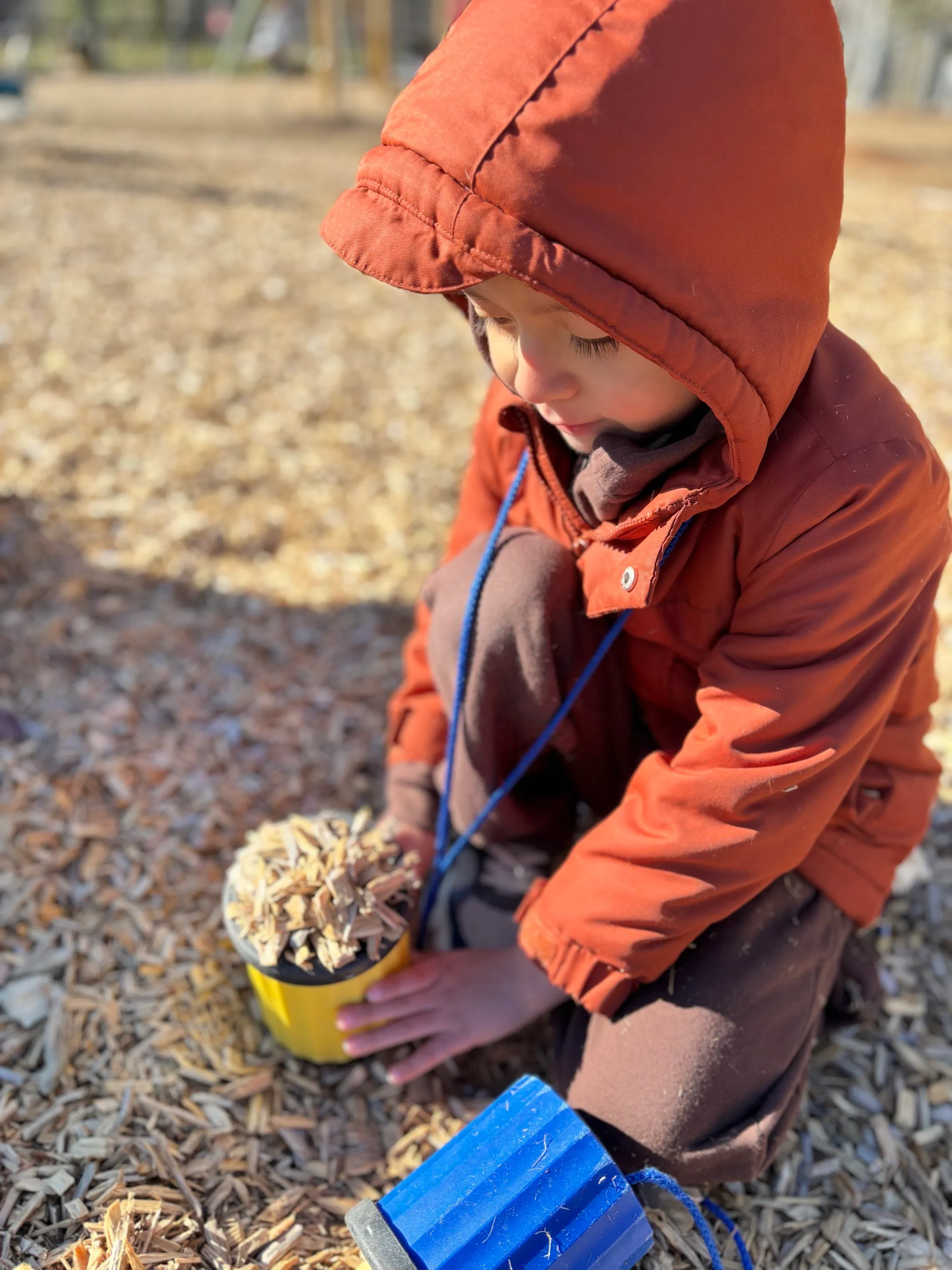 Young child wearing an orange hooded jacket playing with a yellow bucket filled with wood chips on a wood chip-covered ground outdoors.