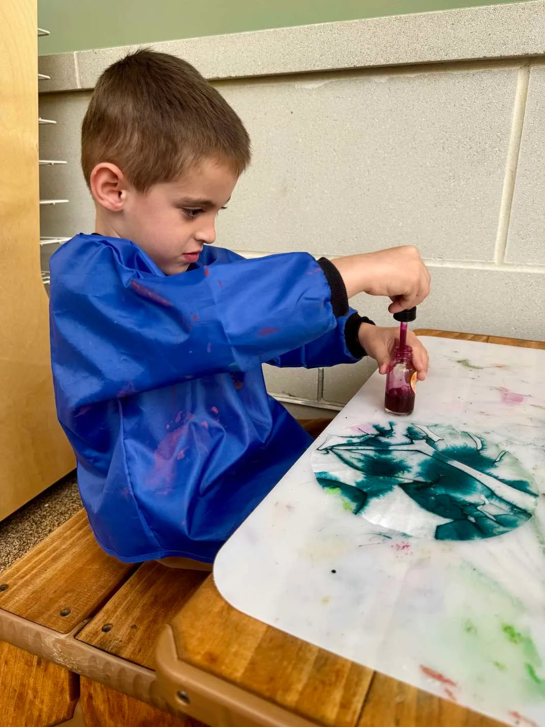 Young boy in a blue smock sitting at a wooden table, concentrating as he squeezes purple liquid from a dropper bottle onto a white surface with green and blue paint stains.