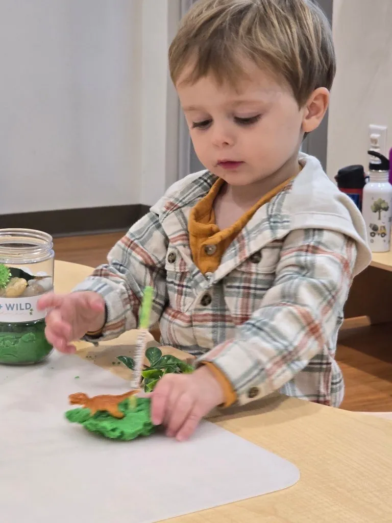Young child in a plaid shirt playing with green playdough and small toy animals at a table.