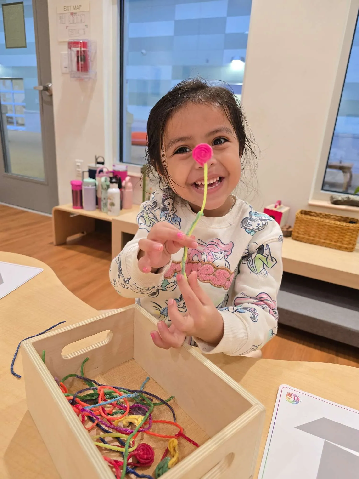 Smiling young girl holding a red yarn flower above a wooden box filled with colorful yarn strings in a playroom.