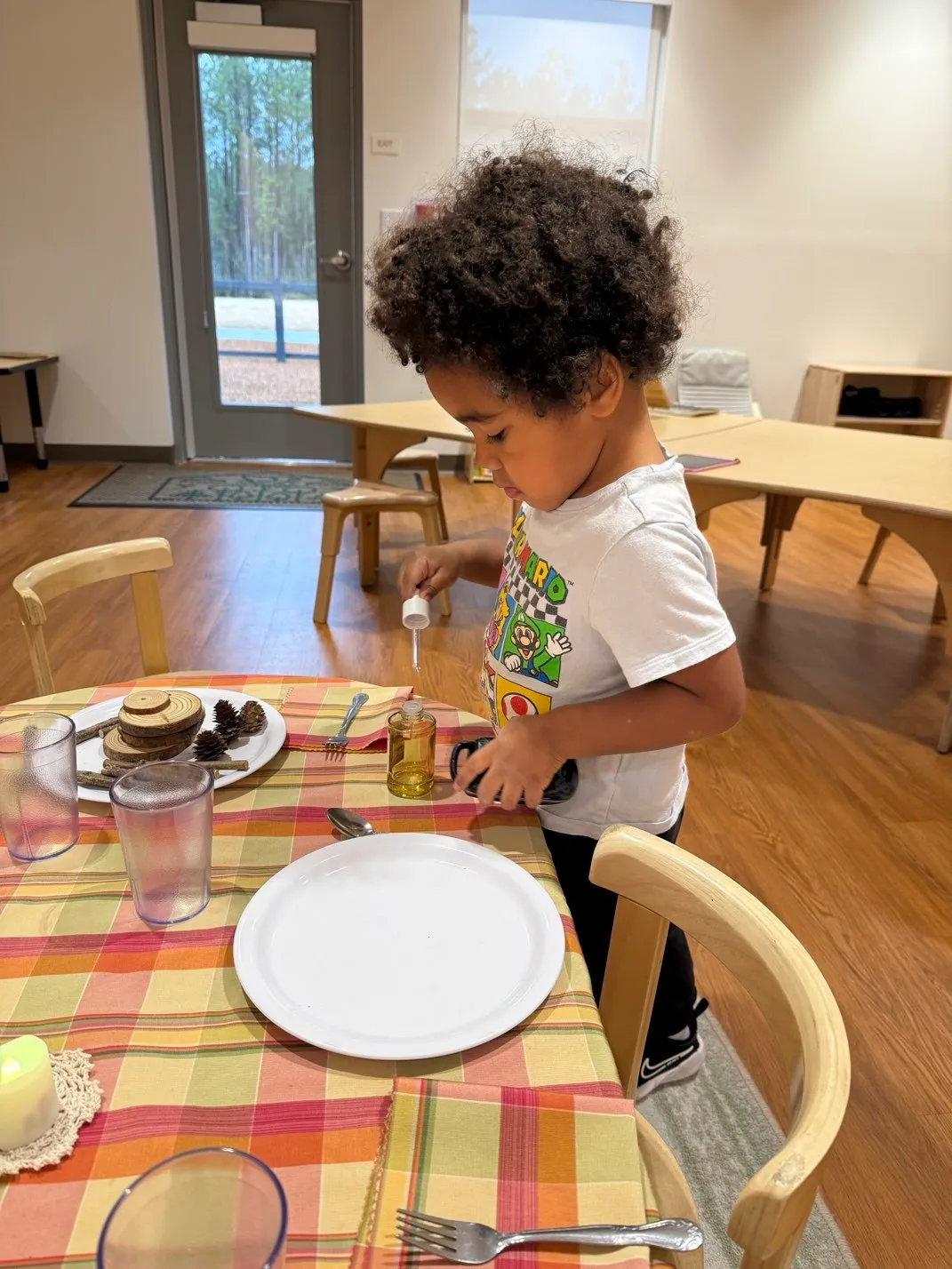 Child with curly hair using a dropper to put liquid from a small bottle on a table with a checkered tablecloth.