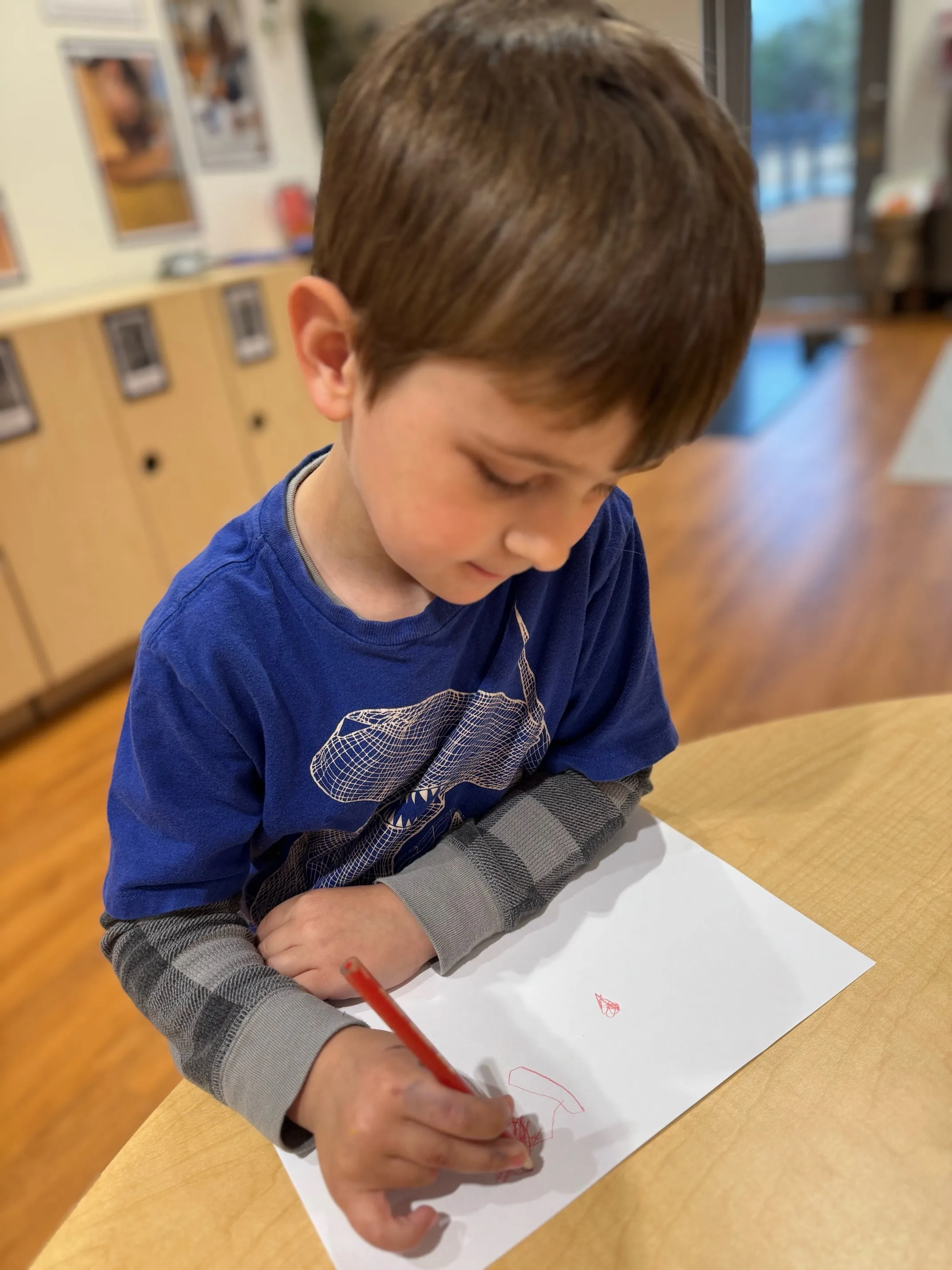 Young boy with brown hair wearing a blue shirt drawing with a red pencil on white paper at a wooden table indoors.