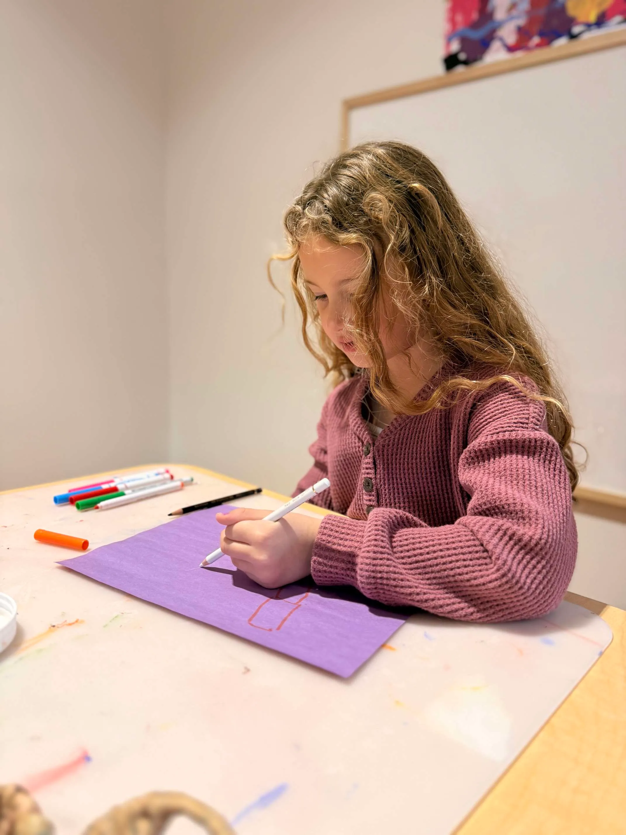 Young girl with curly hair wearing a pink sweater, drawing on purple paper at a table with markers and pencil.