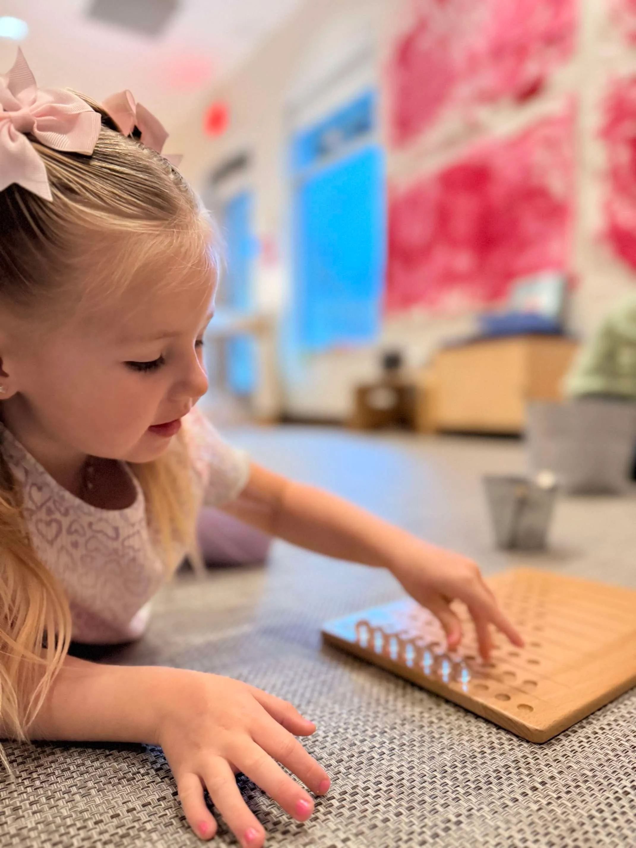 Young girl with pink bows playing with a wooden pegboard toy on a carpeted floor in a bright room.