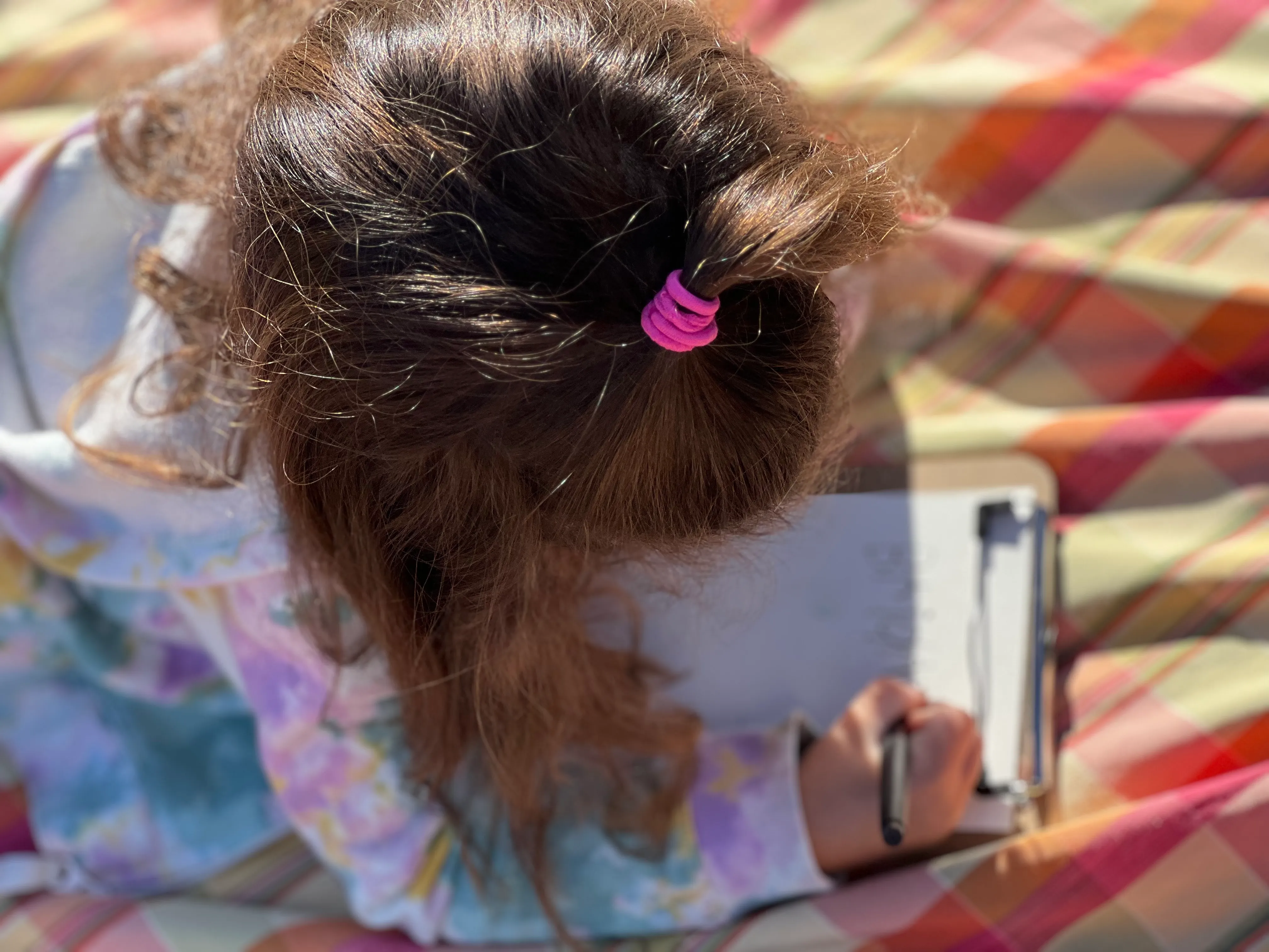 Child with curly hair tied by a pink hairband writing on a clipboard while sitting on a colorful striped blanket.