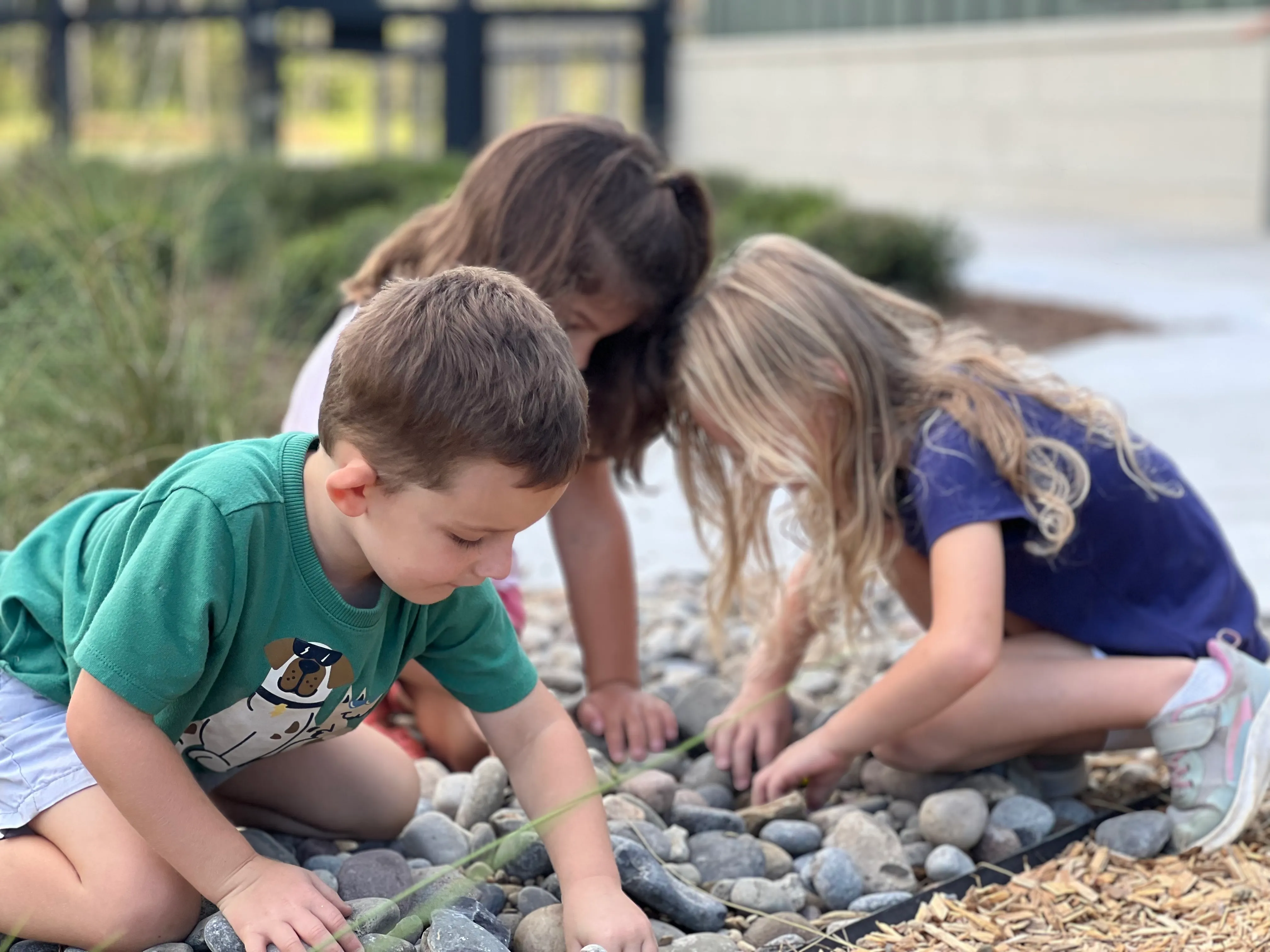 Three children crouched on the ground playing and examining small rocks outdoors.